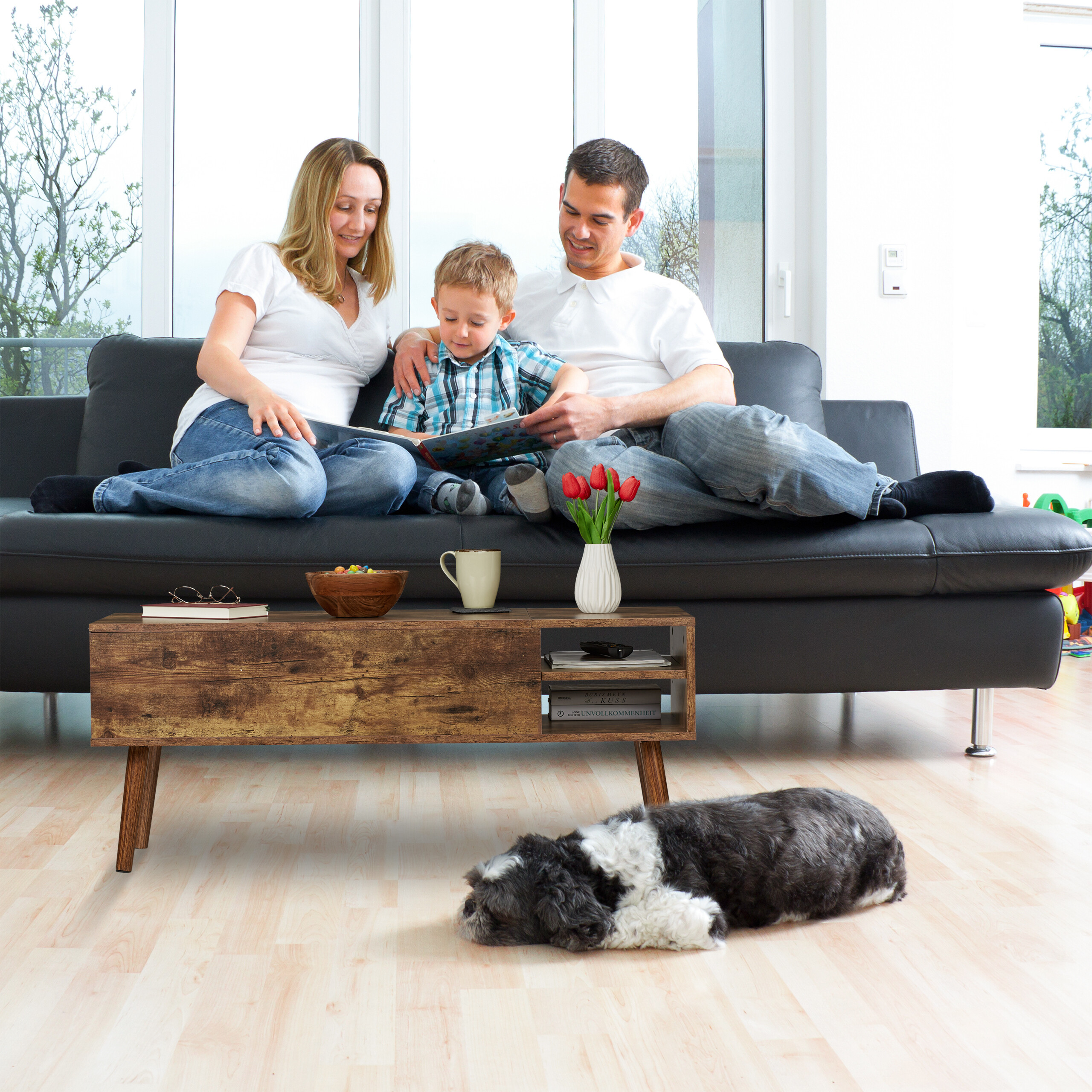 Eine Familie sitzt auf einem schwarzen Sofa und liest mit einem Kind ein Buch, während ein Hund auf dem Holzboden schläft.