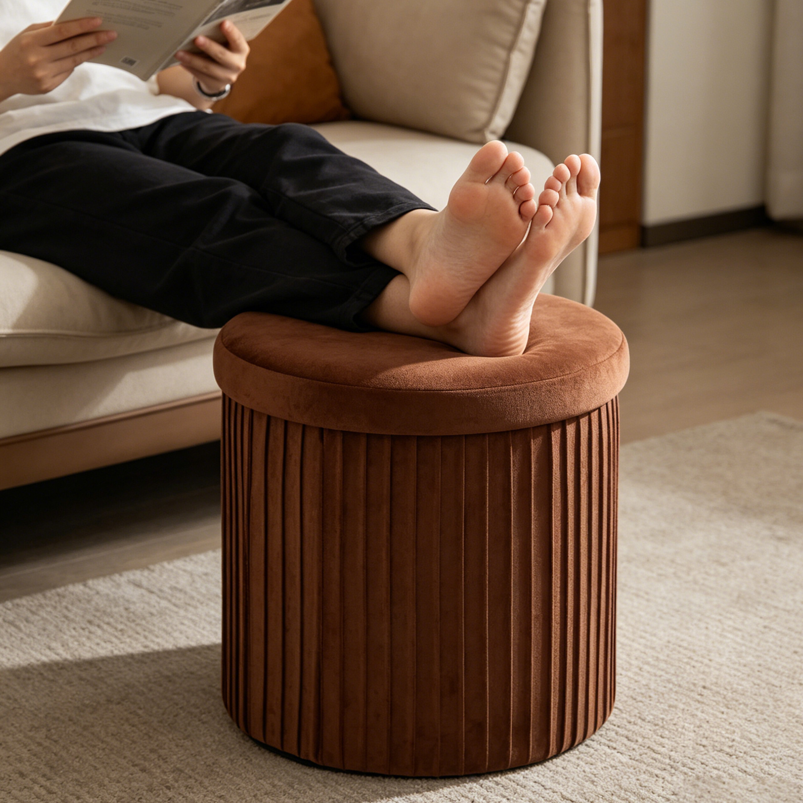 Eine Person liest ein Buch auf einem beige Sofa und legt die nackten Füße auf einen braunen Samt-Hocker.