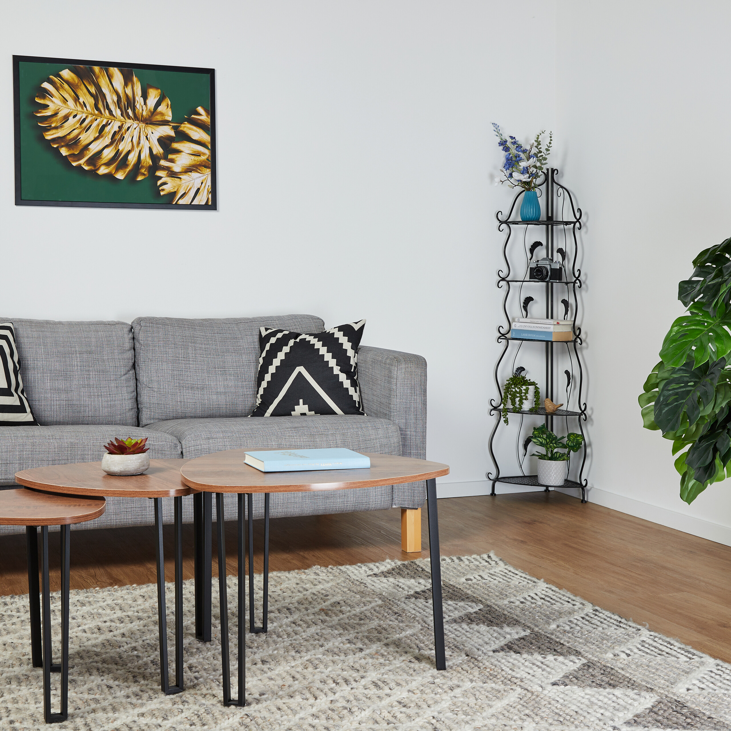Ein Wohnzimmer mit grauem Sofa, Satztischen, einem schwarzen Metallregal und einem Blätter-Kunstdruck.