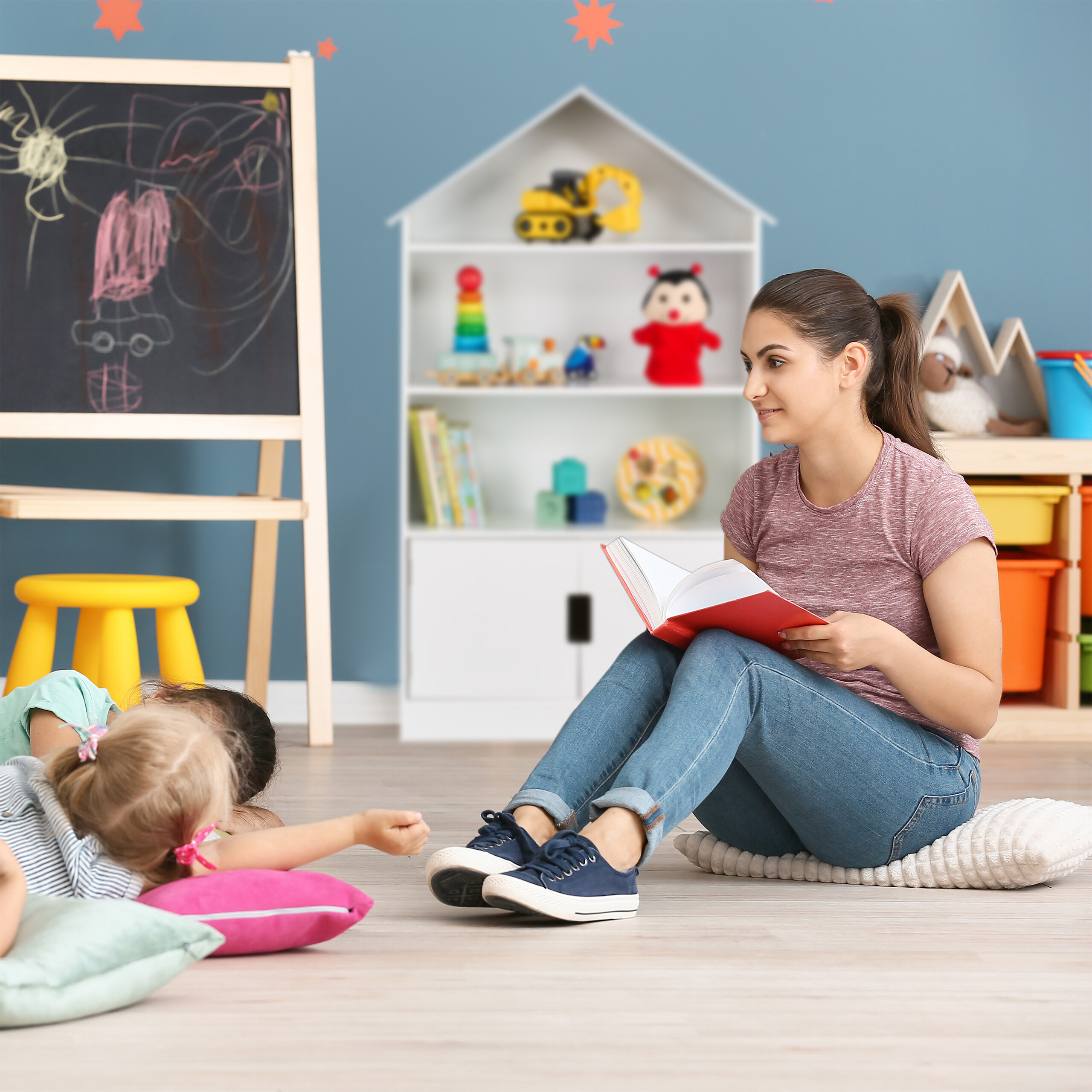 Eine Frau liest Kindern ein Buch vor, die auf Kissen in einem bunten Spielzimmer liegen.