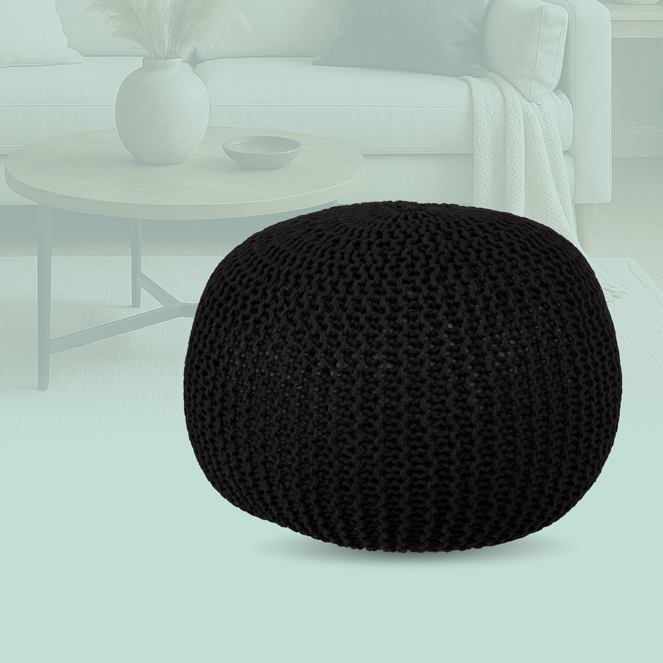 Ein schwarzer Strickpouf steht im Vordergrund vor einem verschwommenen Wohnzimmer.