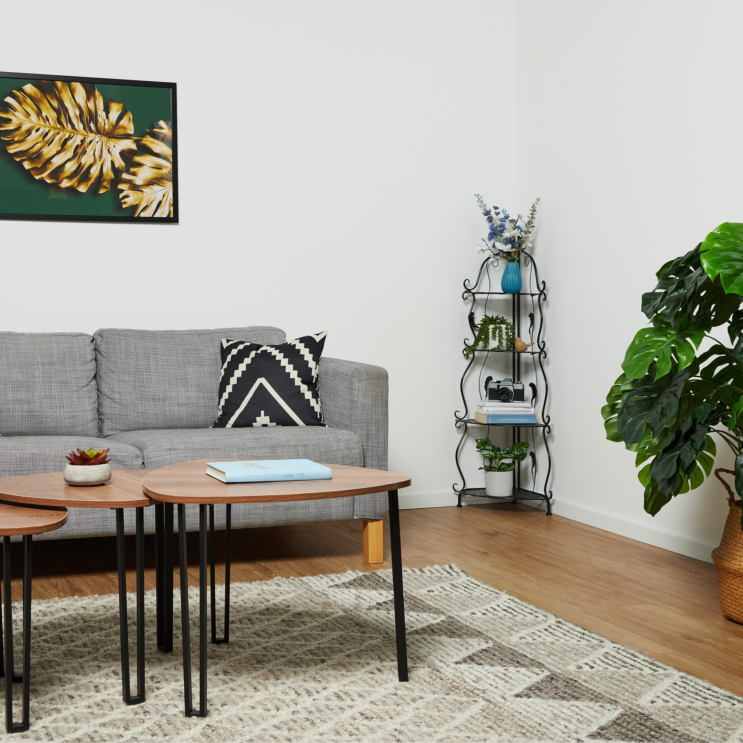 Ein Wohnzimmer mit einem grauen Sofa, Holztischchen, schwarz-weißem Kissen und einem Metall-Eckregal mit Pflanzen.