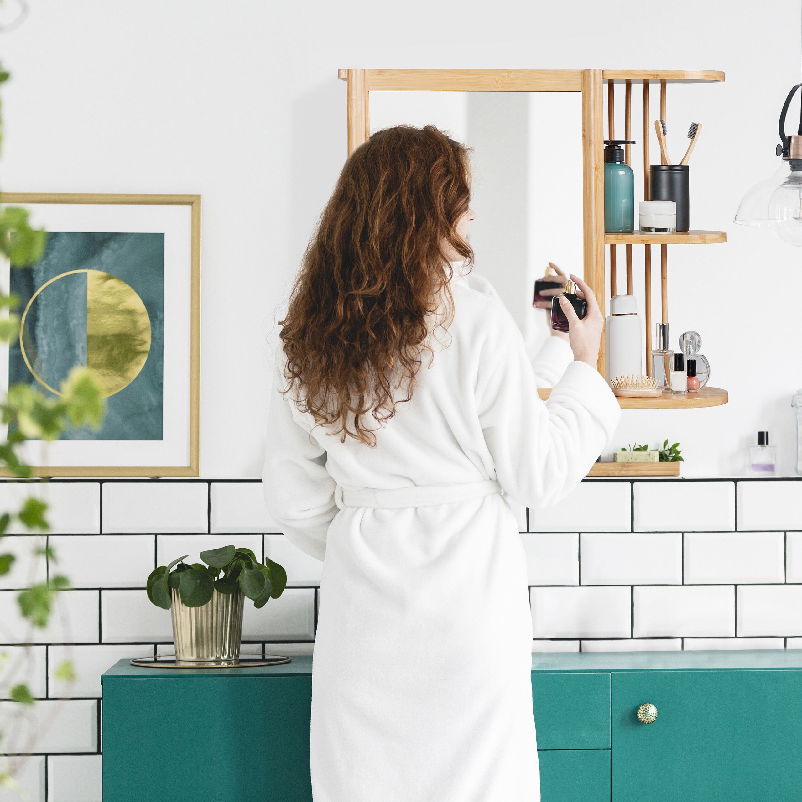 Eine Frau im weißen Bademantel steht vor dem Badezimmerspiegel und hält eine Parfumflasche, Regale mit Kosmetik.