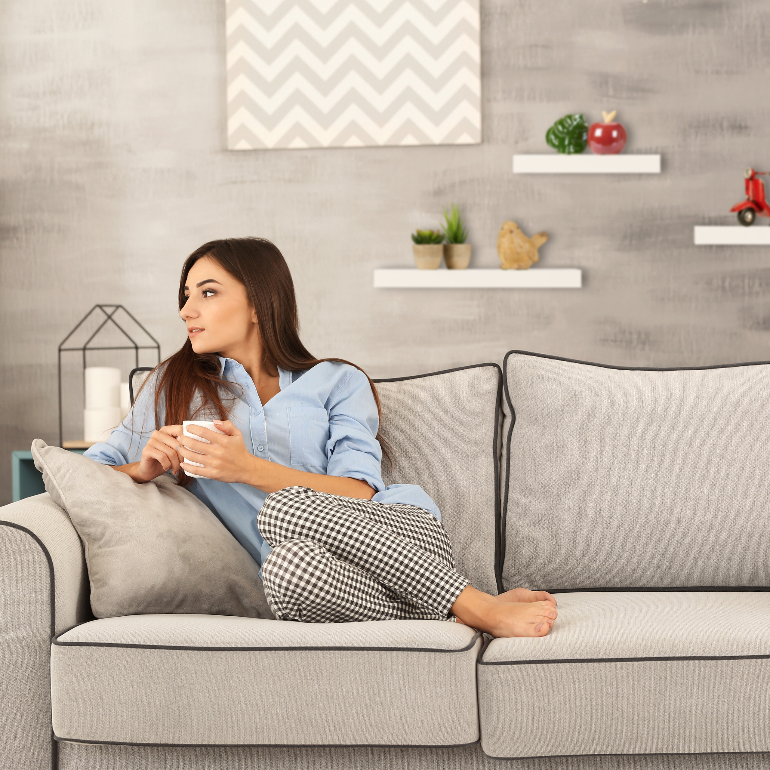 Eine Frau sitzt mit einer Tasse auf einem grauen Sofa in einem modern eingerichteten Wohnzimmer.