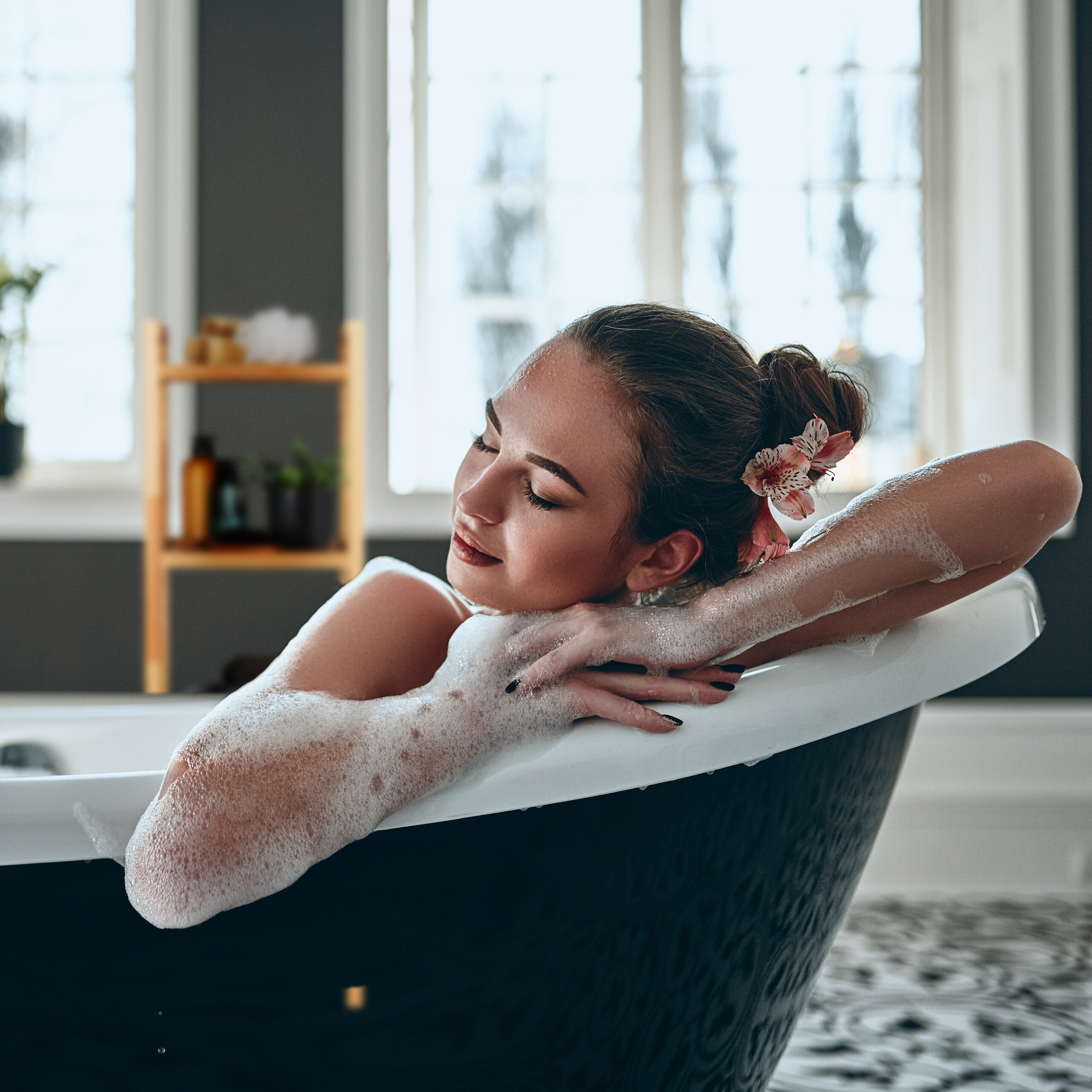 Eine entspannte Frau mit Blumen im Haar ruht sich in einer Badewanne voller Schaum aus.