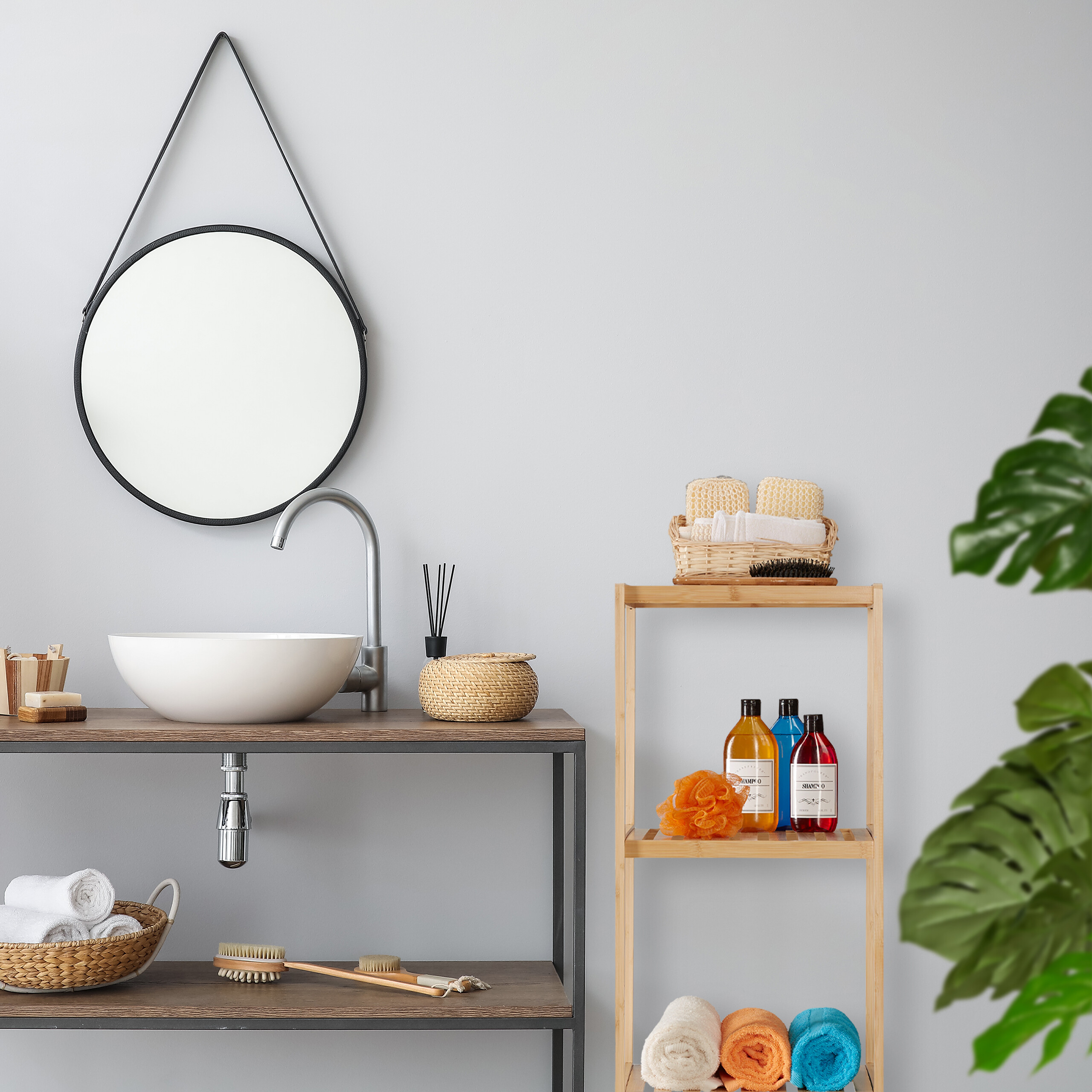 Badezimmer mit Waschbecken, rundem Spiegel, Holzregal voller Badutensilien und einer Monstera-Pflanze im Vordergrund.