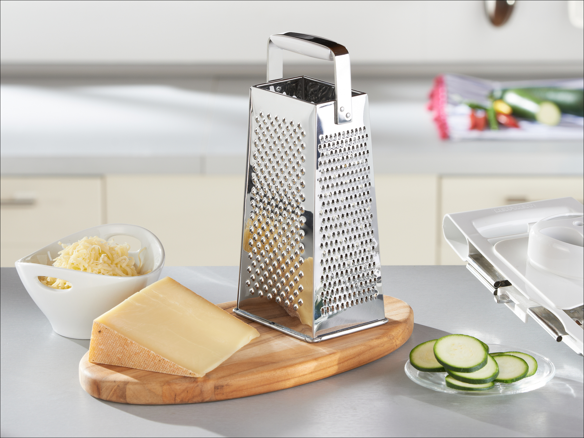 Eine Edelstahl-Vierkantreibe auf einem Holzbrett mit Käse, geriebenem Käse und Gurkenscheiben in einer Küche.