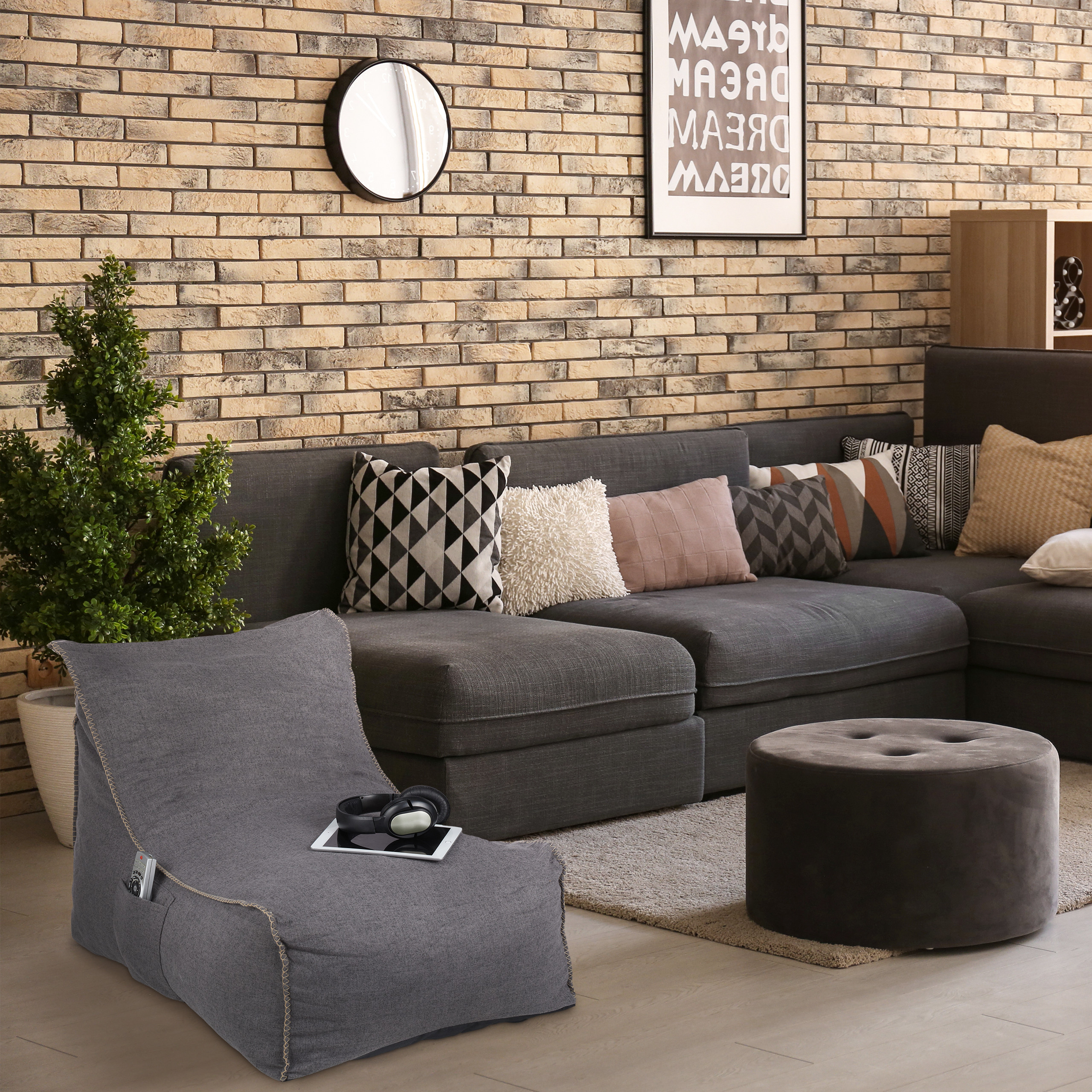 Gemütliches Wohnzimmer mit grauem Sofa, Kissen, Kissenhocker, Pflanze und einer Wanduhr vor einer Ziegelwand.