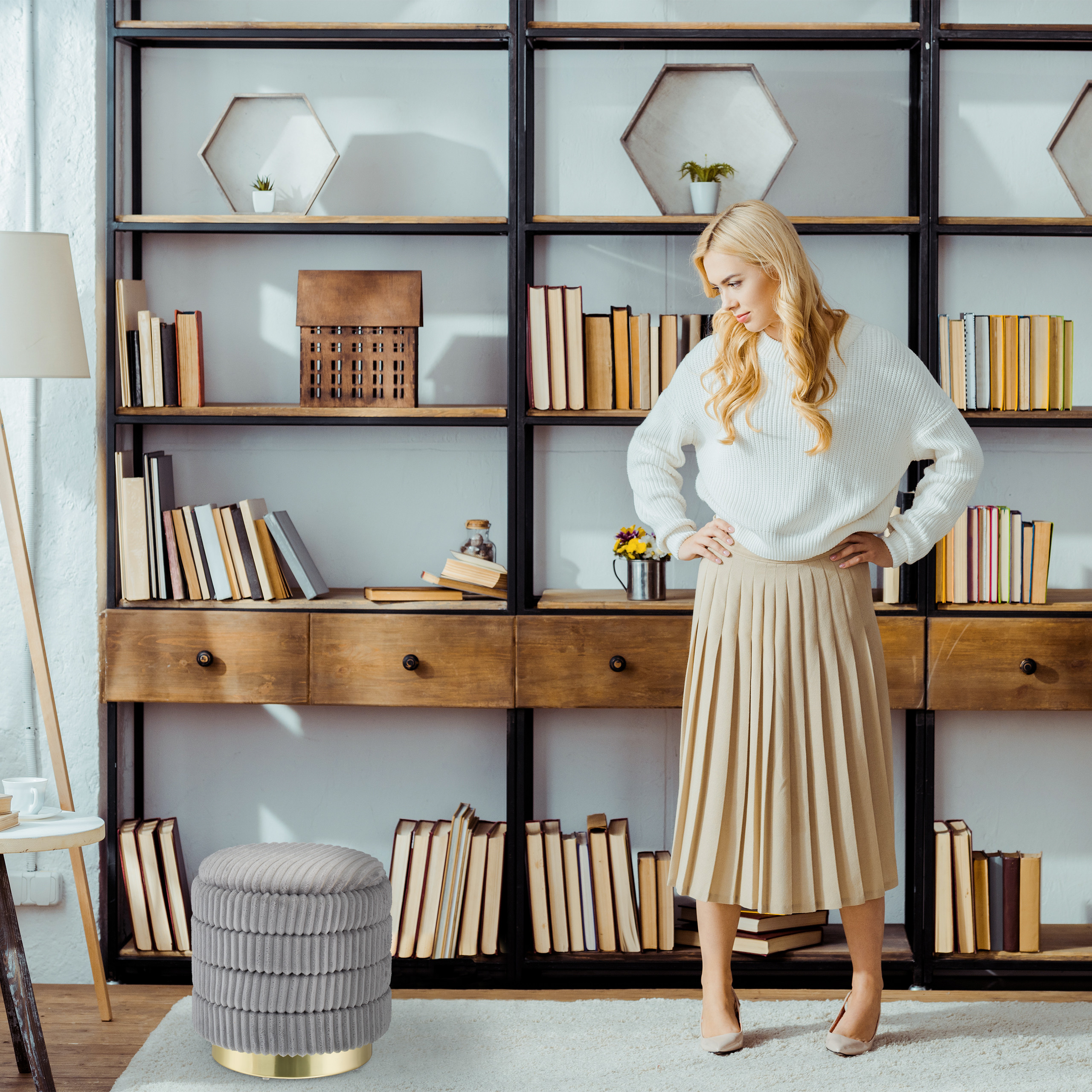 Eine blonde Frau in weißem Pullover und beige Rock steht mit Händen in den Hüften vor einem großen Bücherregal.