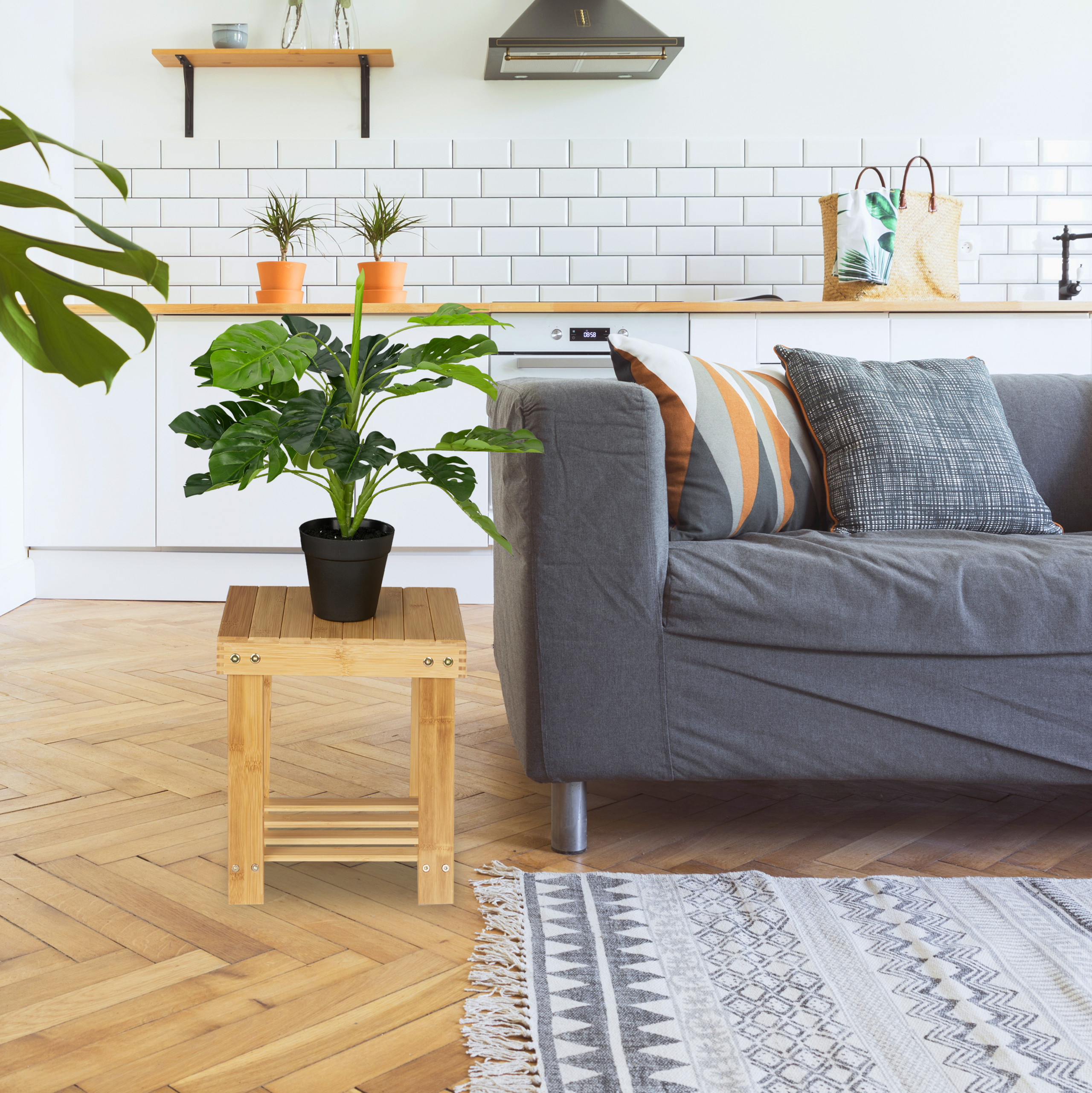 Gemütliches Wohnzimmer mit grauem Sofa, Holztisch, grünen Pflanzen, weißer Küche und gemustertem Teppich.