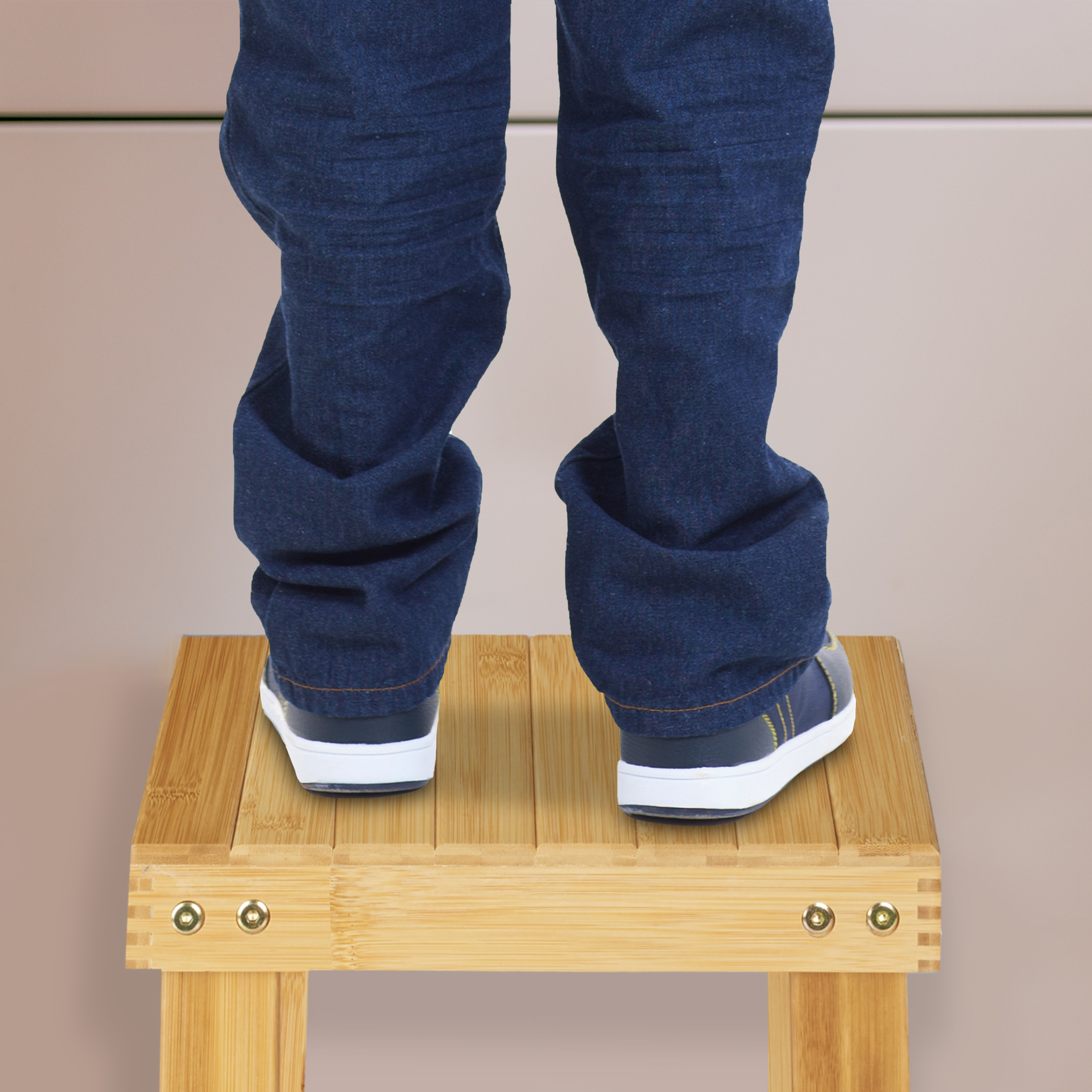 Eine Person in blauen Jeans und Turnschuhen steht auf einem kleinen Holzhocker vor einer hellen Wand.