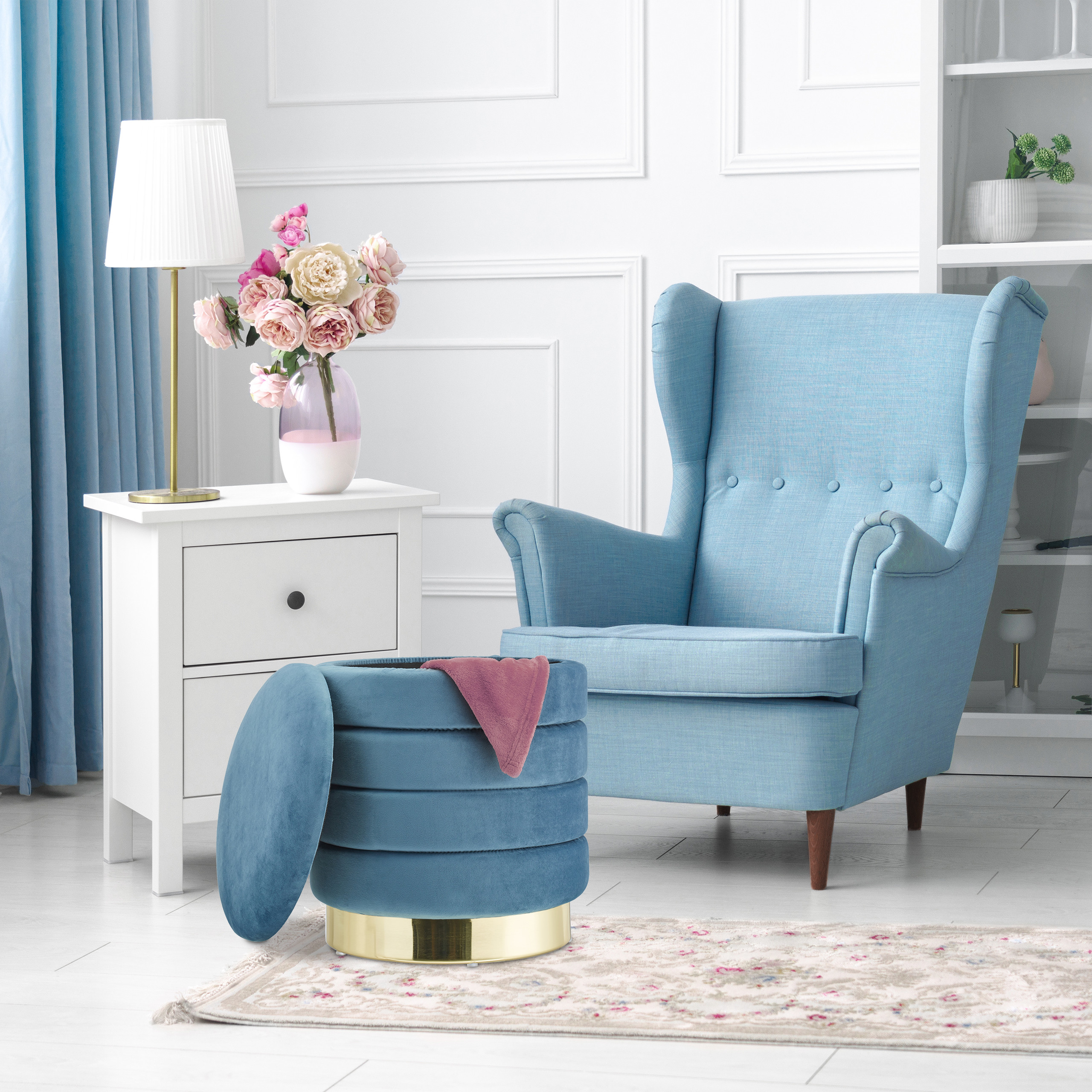 Ein blauer Ohrensessel, ein blauer Hocker mit Goldrand, ein weißer Nachttisch mit Lampe und Blumen stehen in einem hellen Raum.