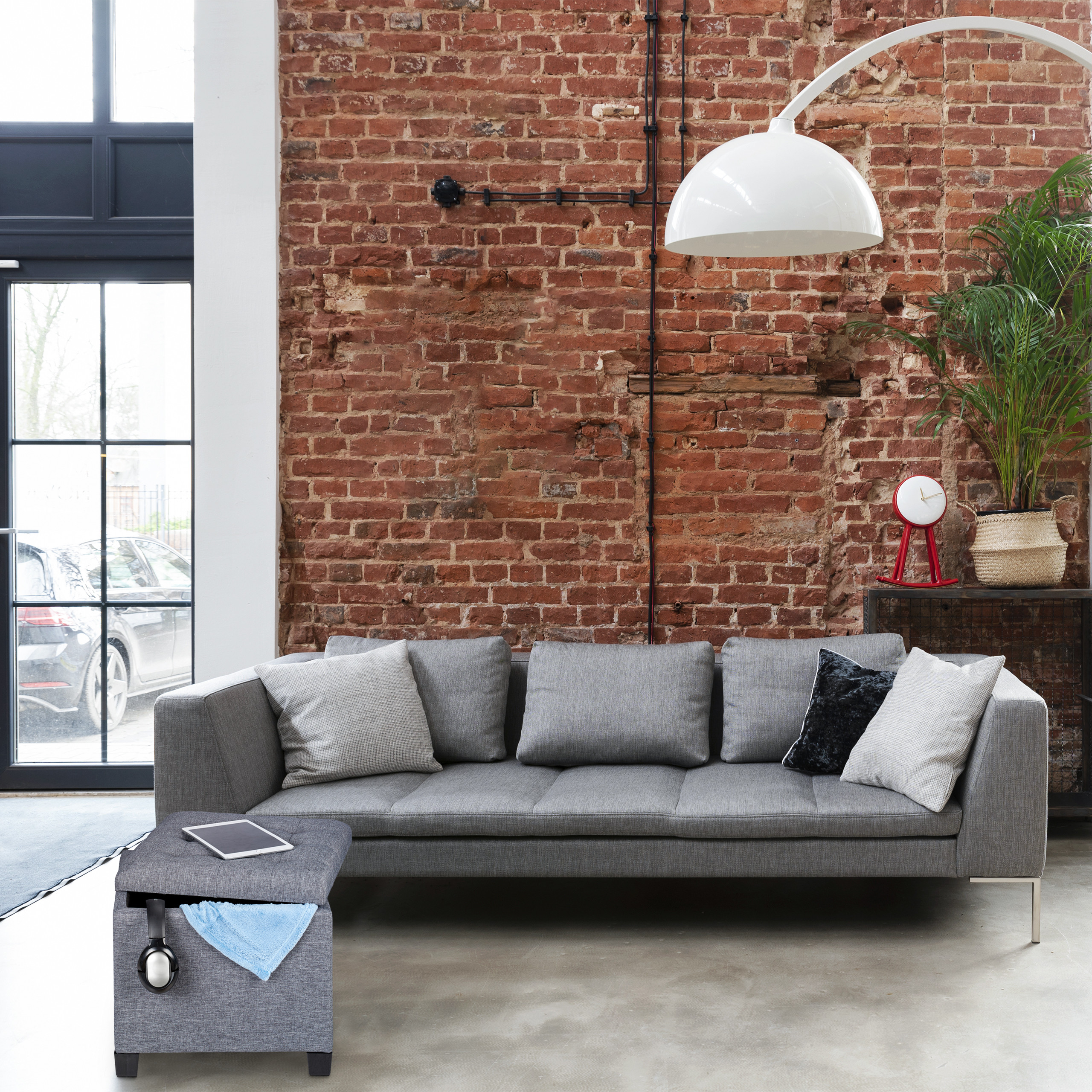 Graues Sofa vor roter Backsteinwand mit großer weißer Lampe, grünem Pflanzen und Fußbank mit Tablet.
