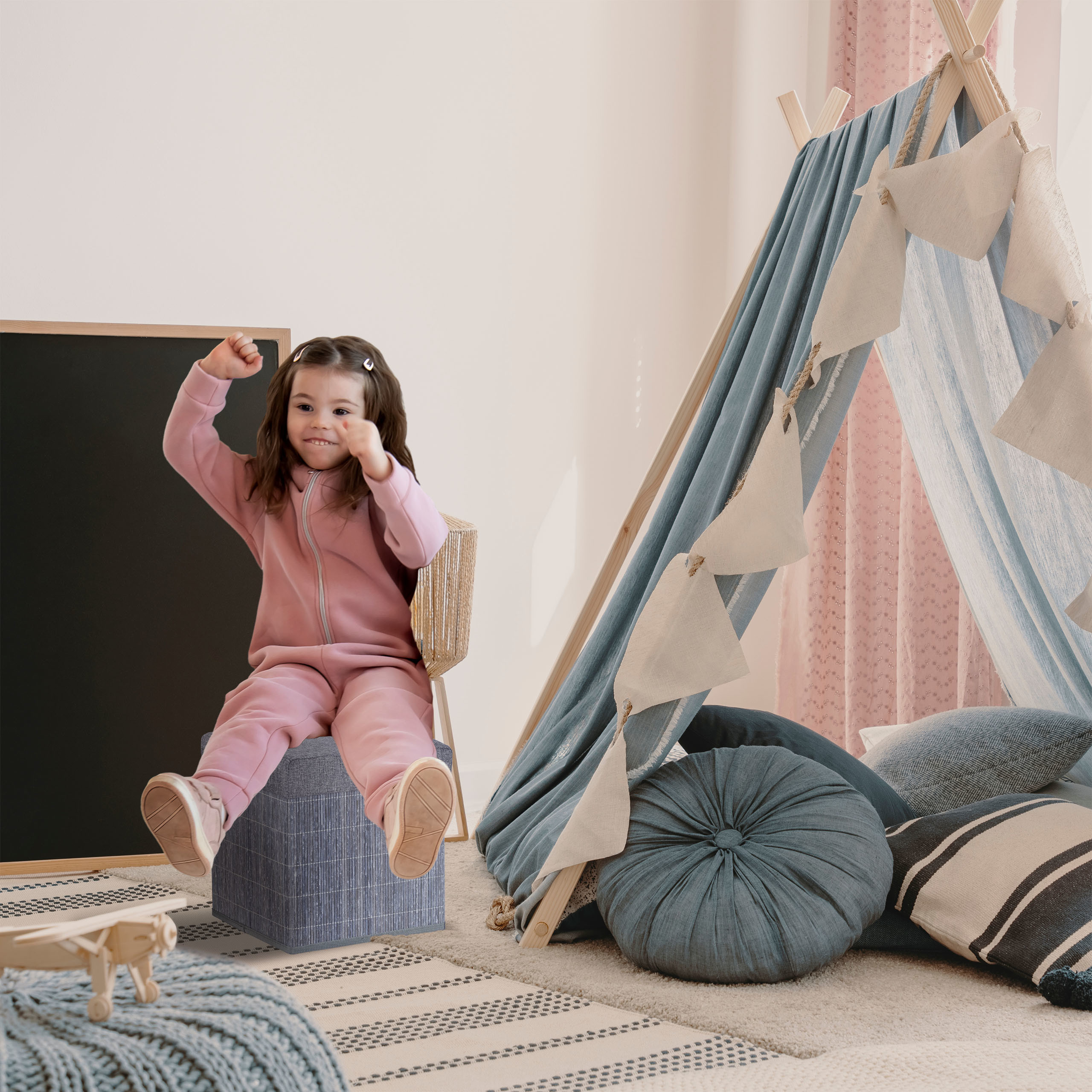 Ein lächelndes Kleinkind in Rosa springt auf einen blauen Hocker in einem Spielzimmer mit Tipi-Zelt.