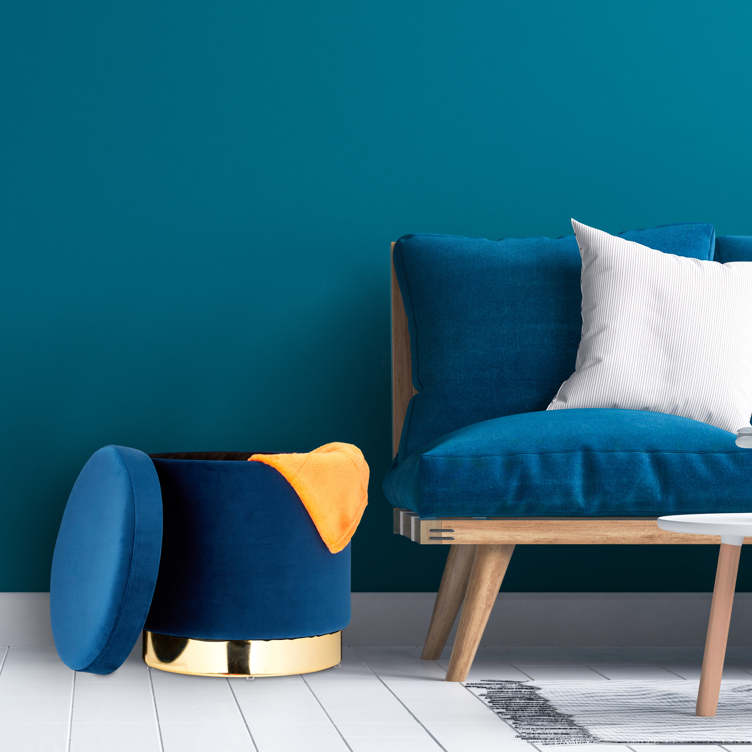 Ein blaues Samtsofa mit weißem Kissen, daneben ein blauer Hocker mit gelber Decke und ein kleiner Tisch vor einer blauen Wand.