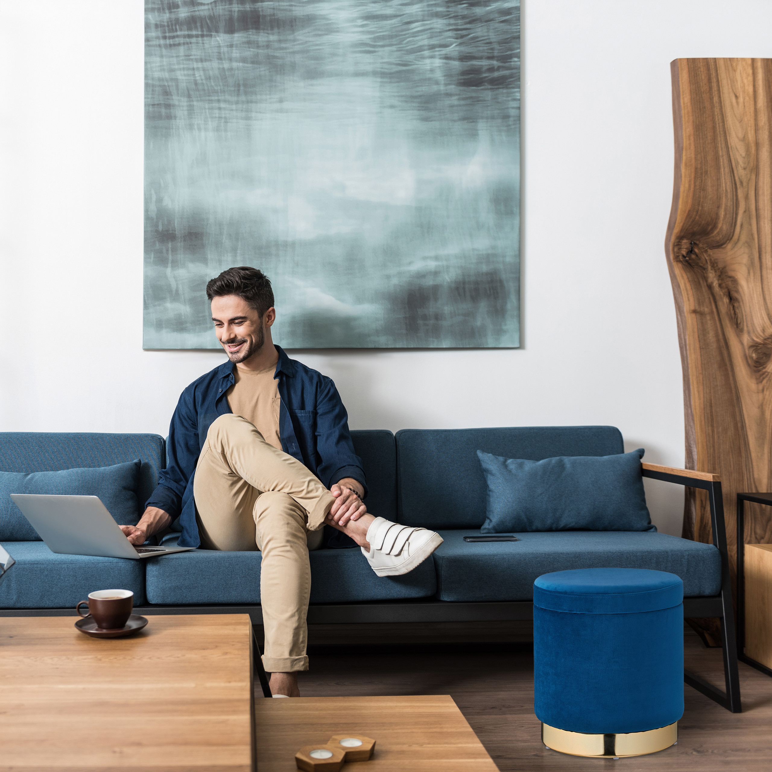 Ein lächelnder Mann sitzt mit gekreuzten Beinen auf einem blauen Sofa und arbeitet an einem Laptop.