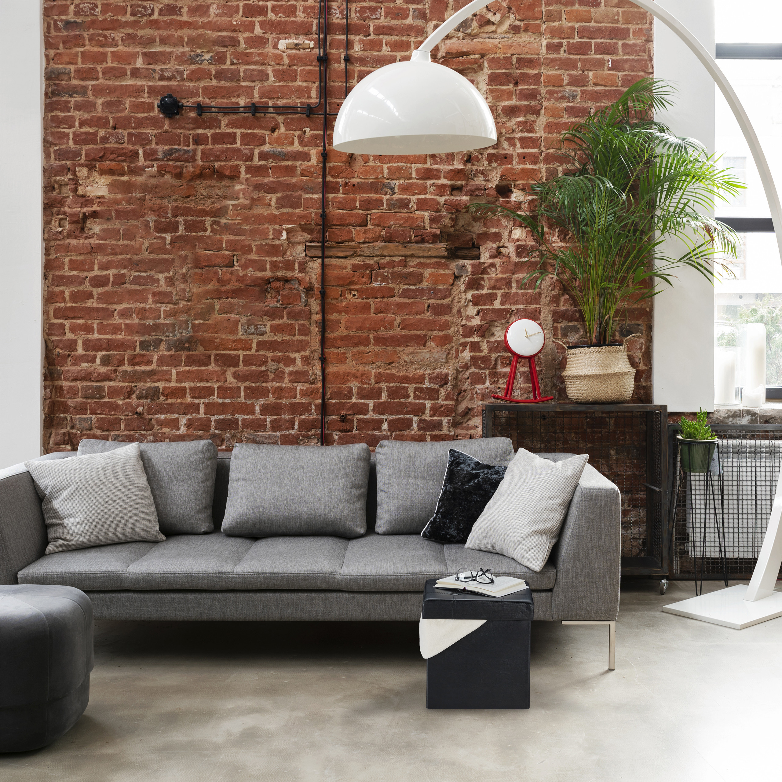 Ein graues Sofa steht vor einer roten Ziegelwand, mit Kissen, einer grossen Bogenlampe und einer Pflanze.