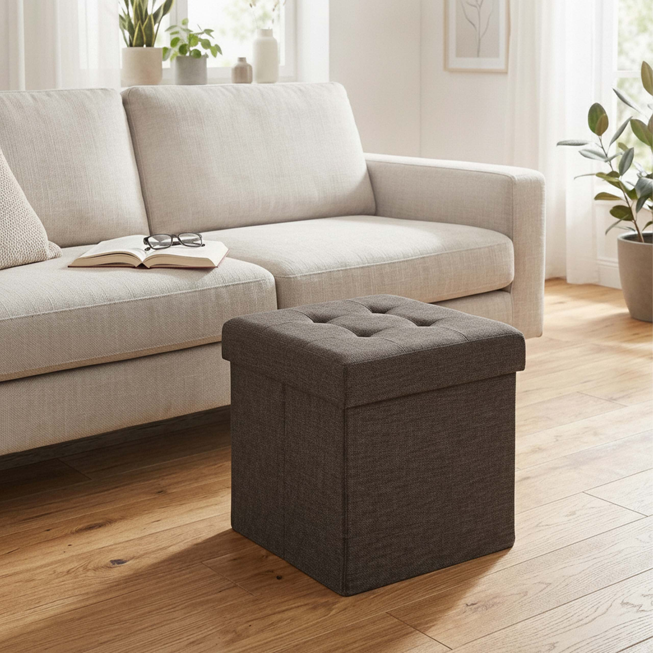 Ein dunkelgrauer Stoffhocker steht auf dem Holzfußboden vor einem hellen Sofa in einem hellen Wohnzimmer.