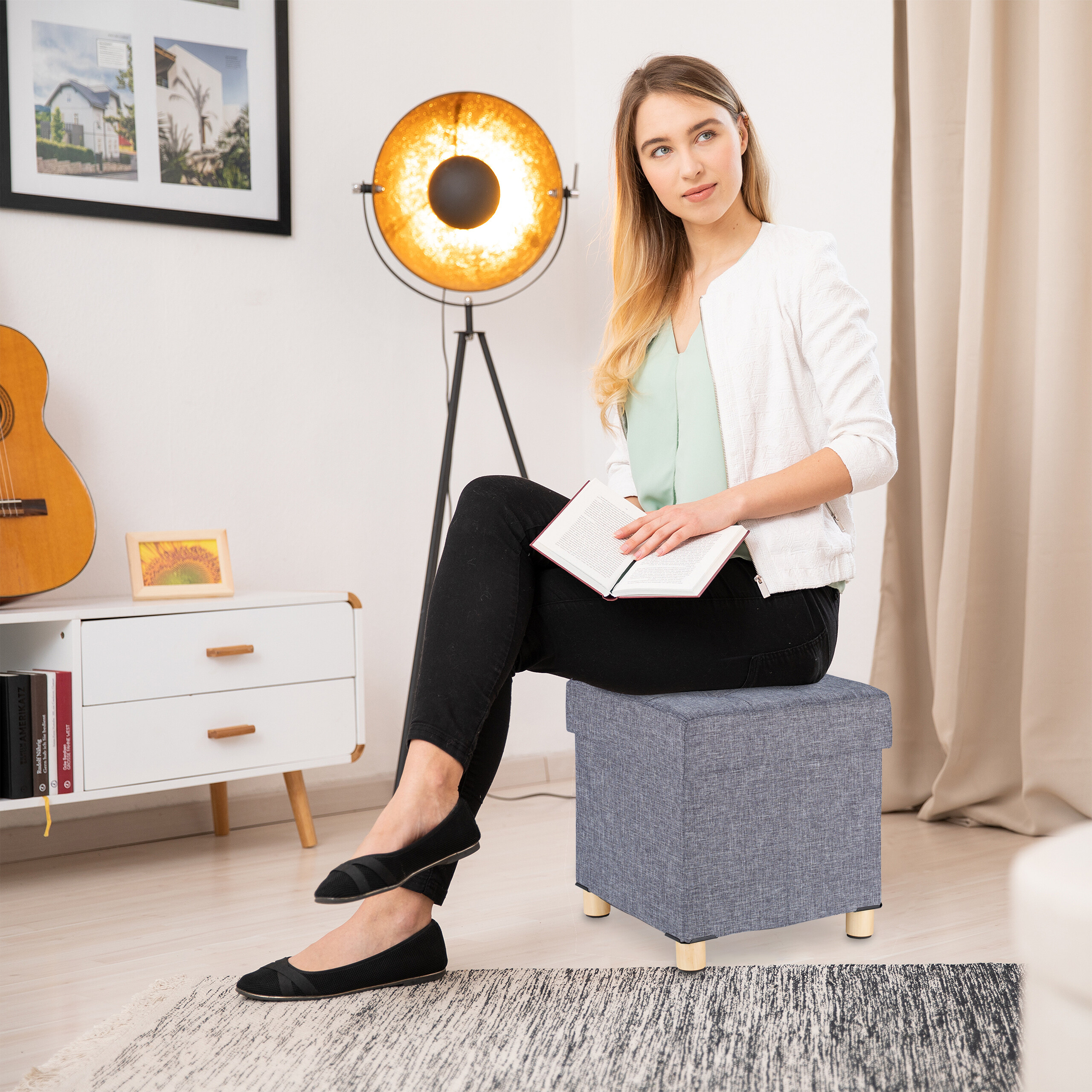 Eine Frau mit einem Buch sitzt auf einem blauen Hocker in einem hellen Wohnzimmer mit Lampe und Gitarre.