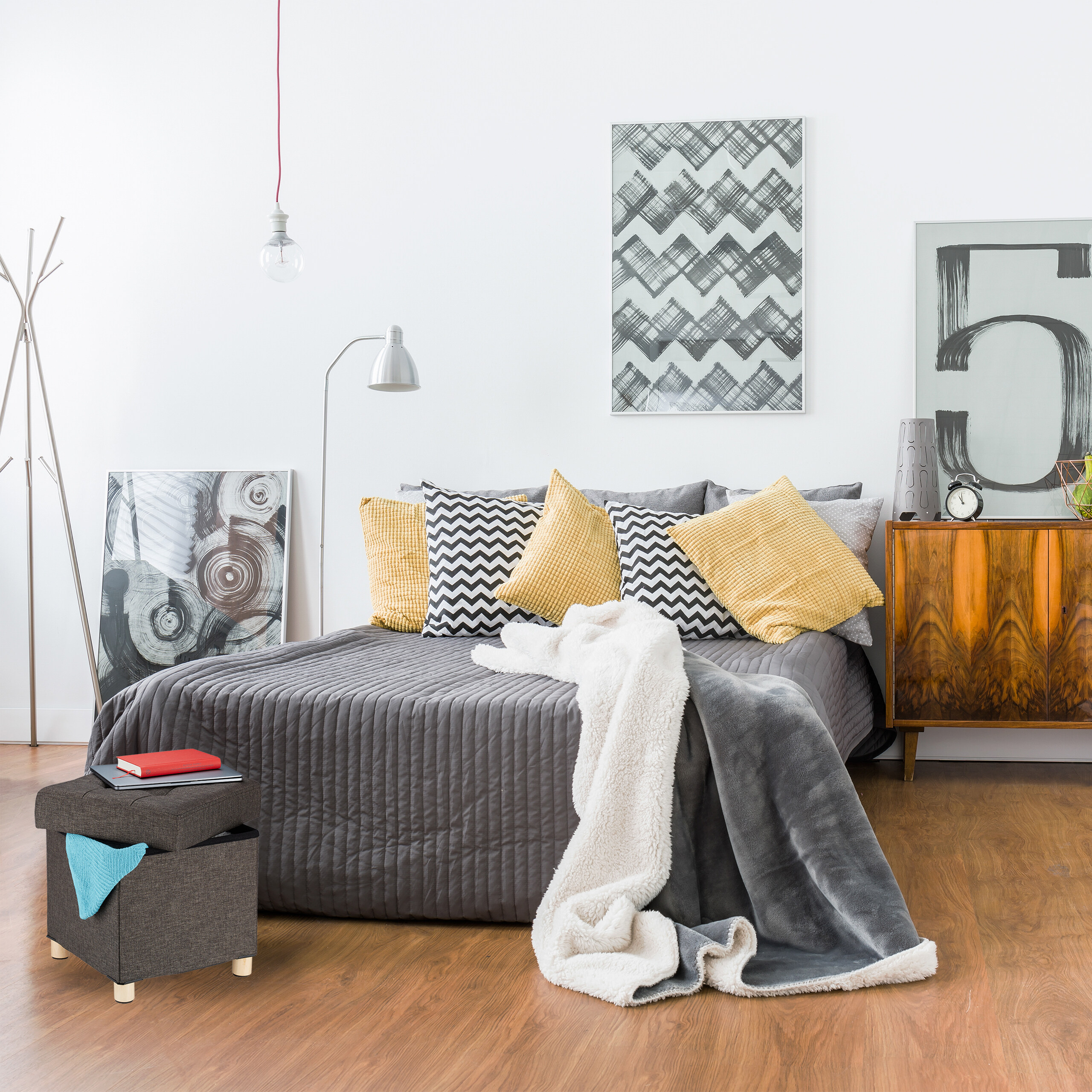 Ein modernes Schlafzimmer mit grauem Bett, gelben Kissen, Wandkunst und einem Holzkasten.