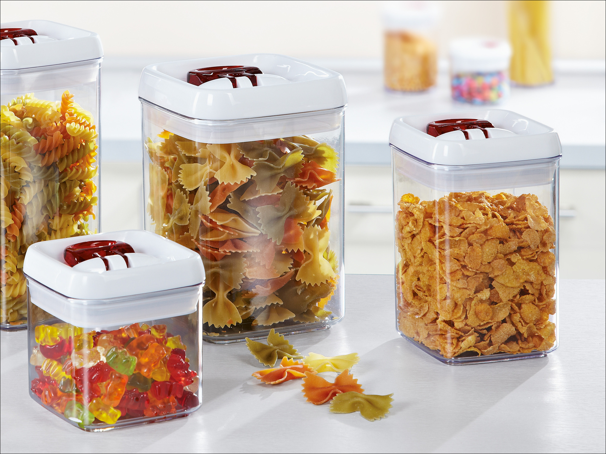 Vier klare Vorratsdosen mit weißem Deckel auf einer Theke, gefüllt mit Nudeln, Cornflakes und Gummibärchen.