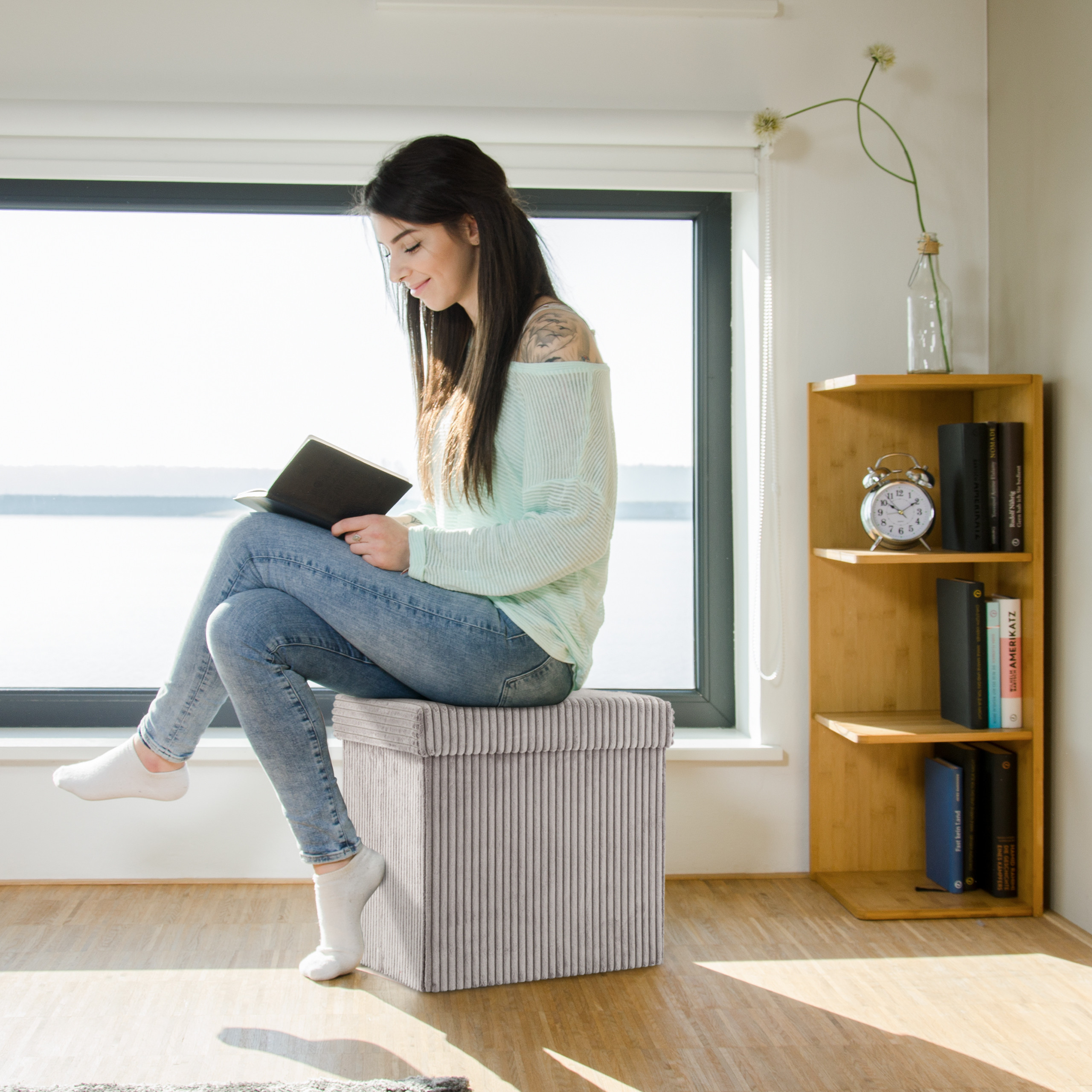Eine Frau sitzt auf einem grauen Hocker vor einem Fenster und liest ein Buch.