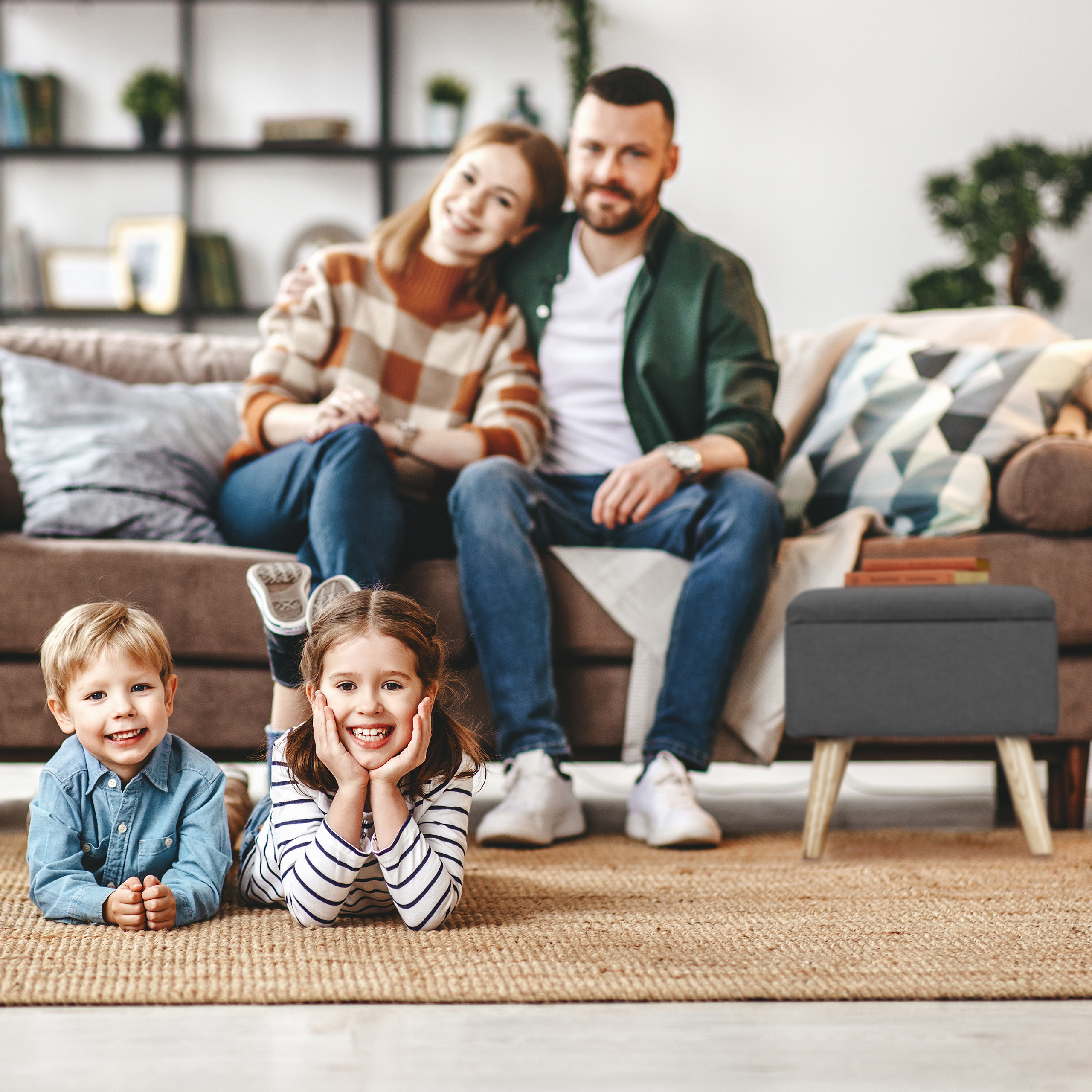 Zwei glückliche Kinder liegen auf dem Teppich, während ihre Eltern im Hintergrund auf dem Sofa sitzen.