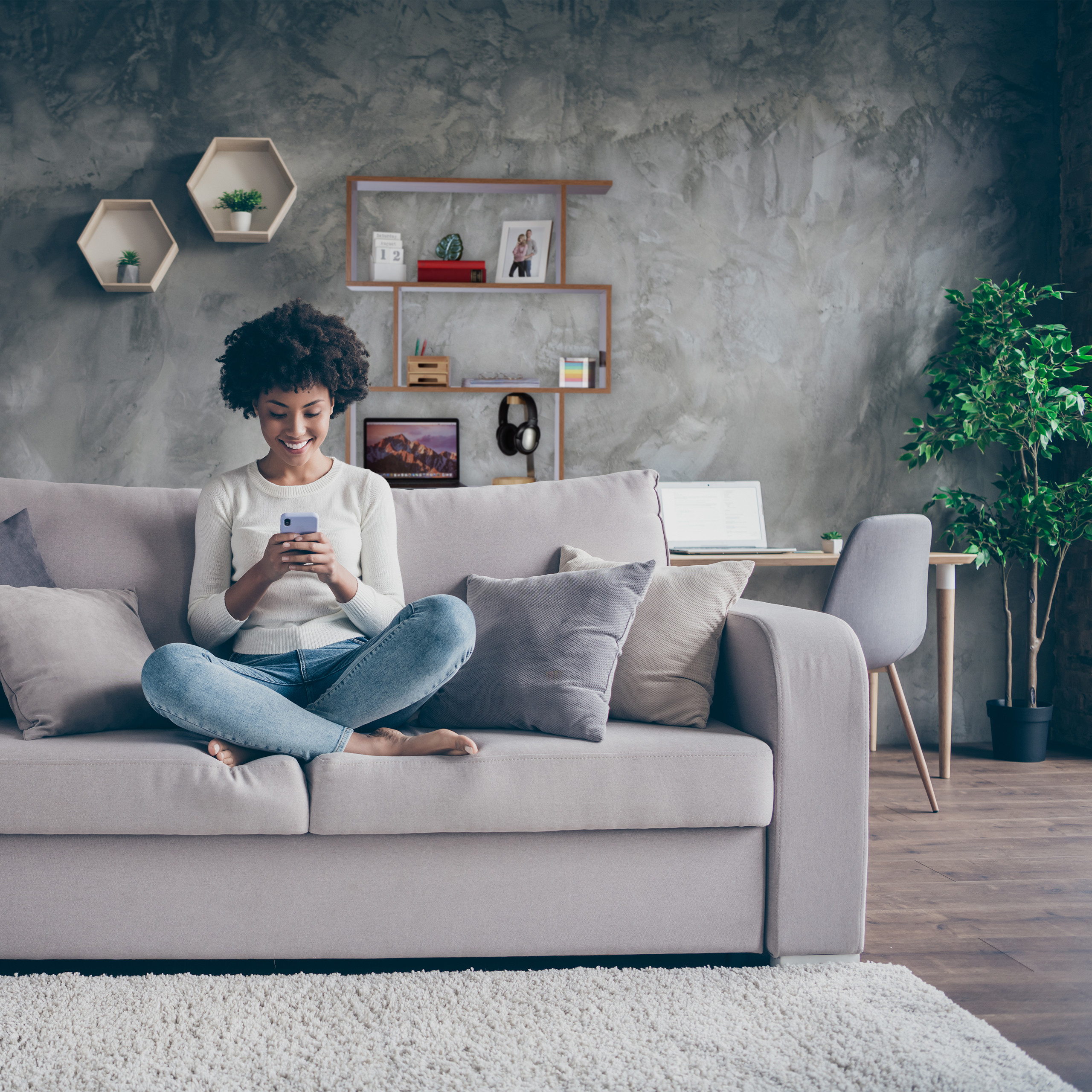 Eine Frau sitzt auf einem grauen Sofa und lächelt auf ihr Smartphone.