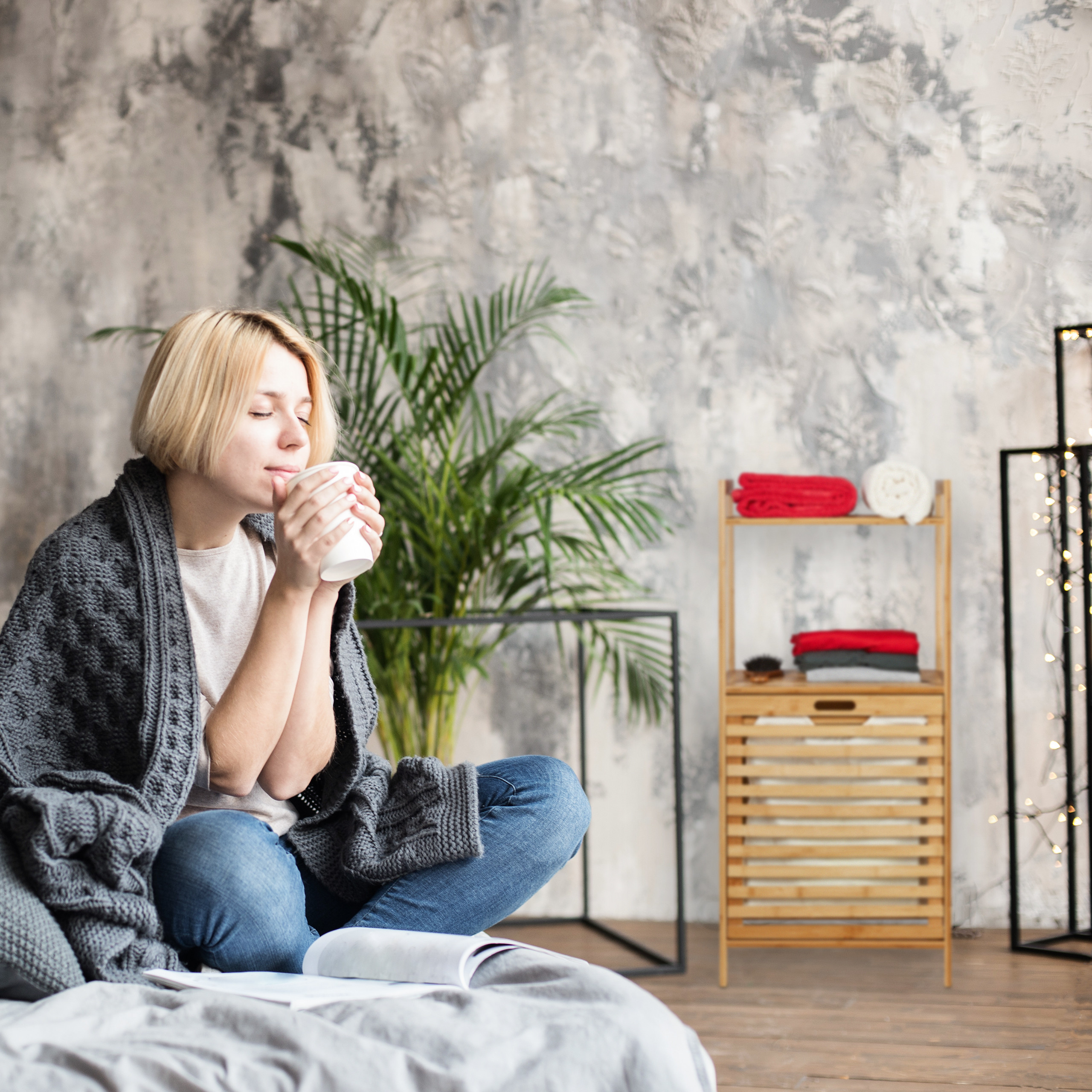 Blonde Frau mit grauer Decke sitzt auf dem Bett und hält eine Tasse.