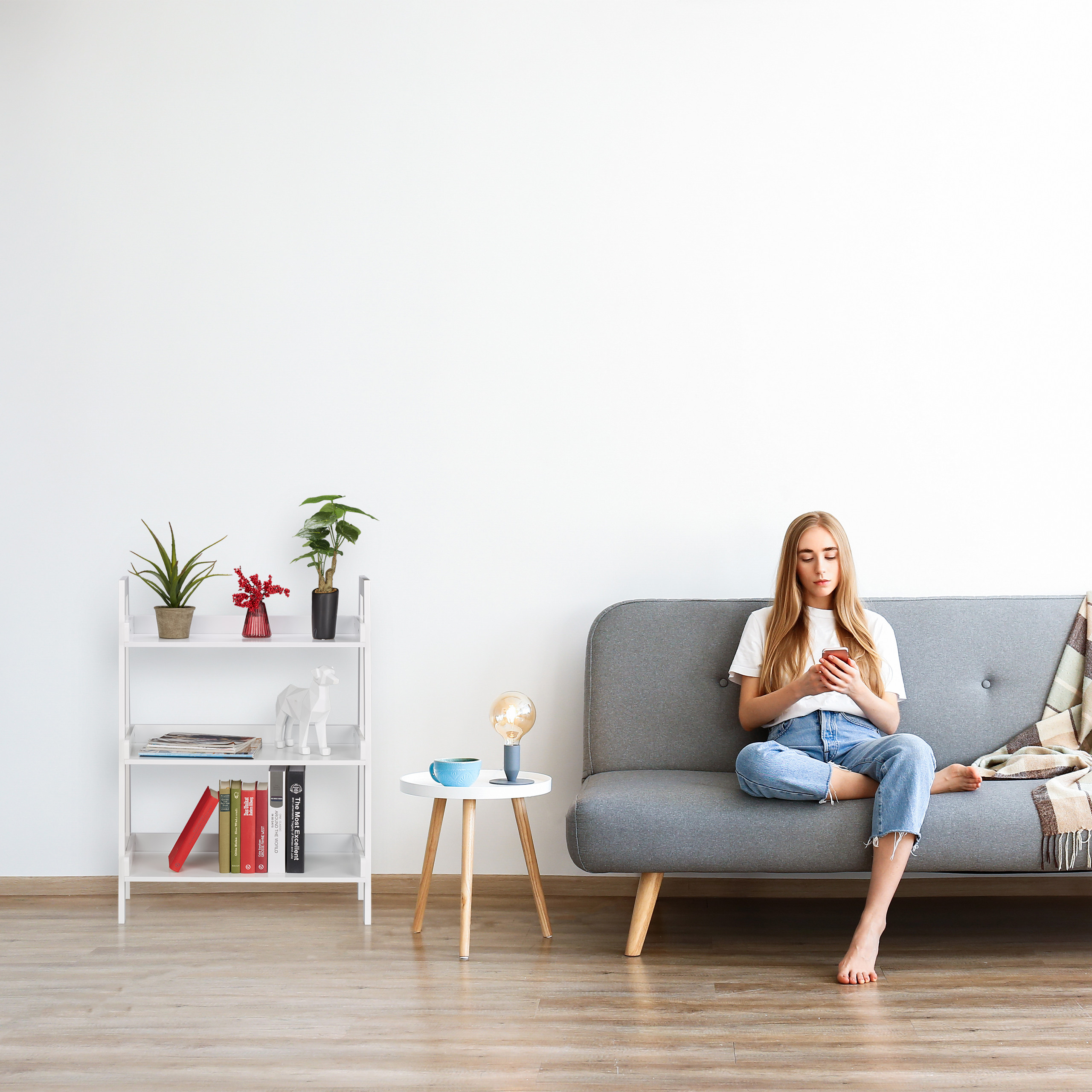 Eine junge Frau sitzt auf einem grauen Sofa und schaut auf ihr Smartphone, neben einem Regal und Tisch.