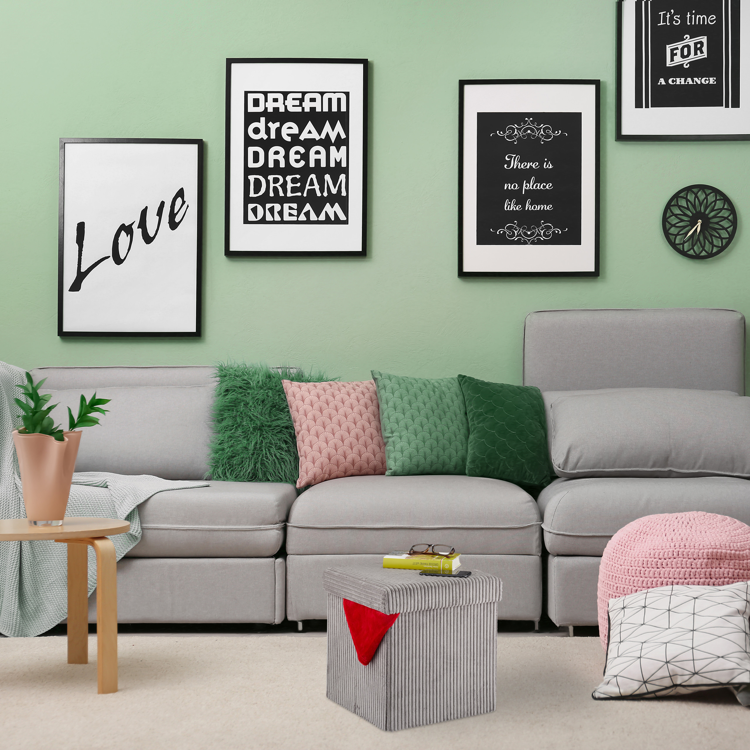 Ein graues Sofa an einer grünen Wand mit gerahmten Bildern, einem kleinen Tisch mit Pflanze und Hocker.