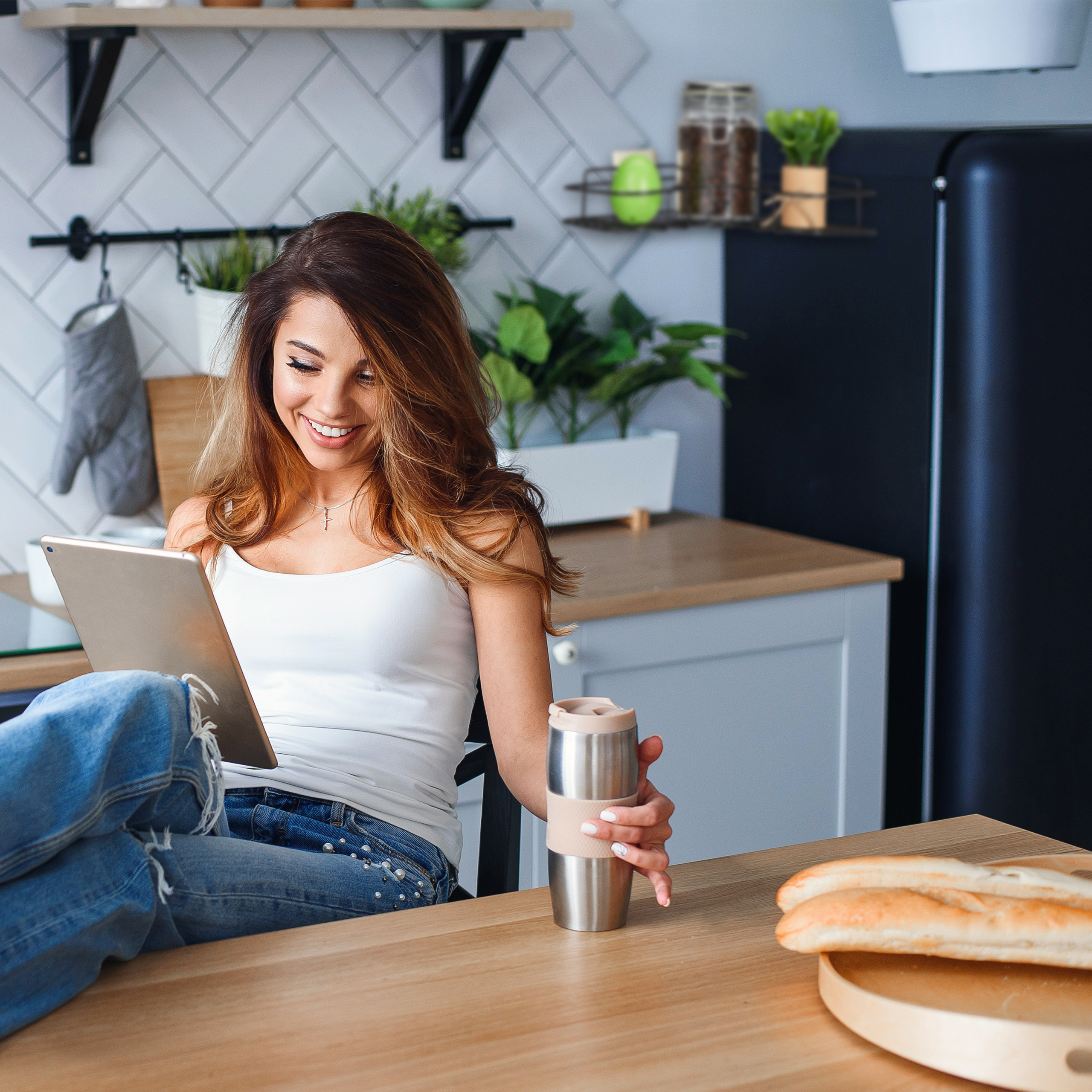 Eine lächelnde Frau in Jeans und weißem Top sitzt in einer Küche, nutzt ein Tablet und hält einen Becher.