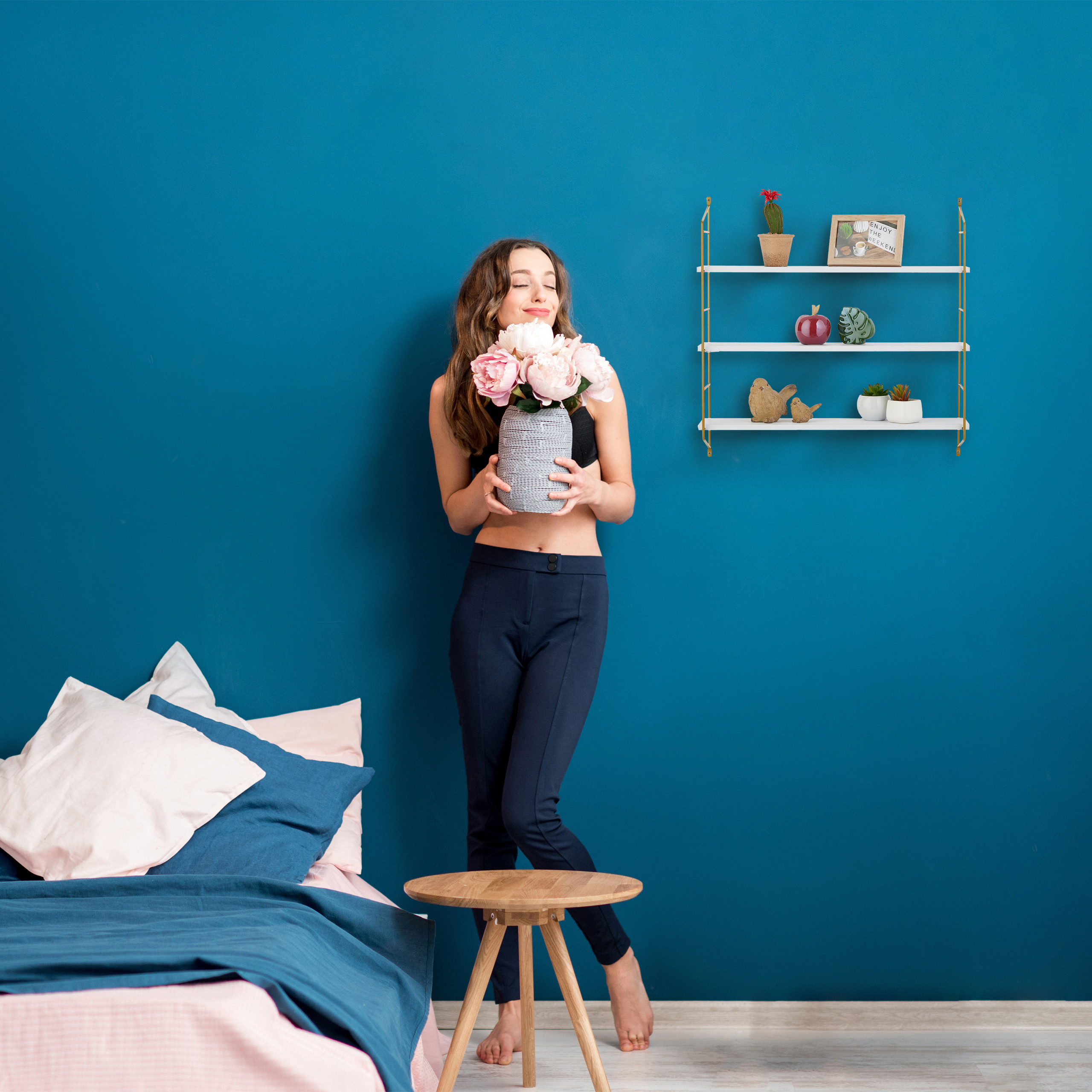 Eine Frau hält eine Vase mit rosa Blumen in einem blauen Zimmer mit Bett und Wandregal.