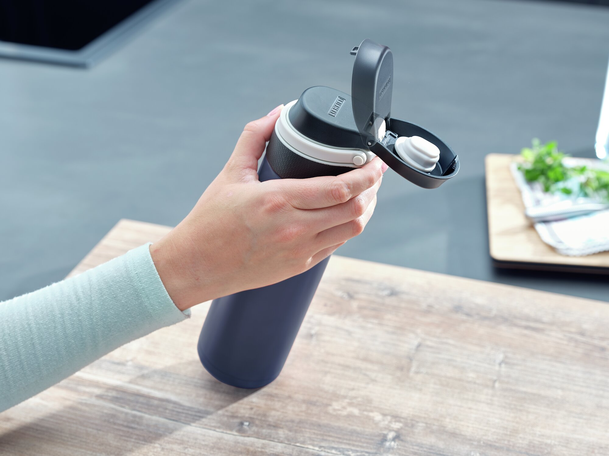 Eine Hand hält eine geöffnete, dunkelblaue Thermosflasche auf einem Holztisch.