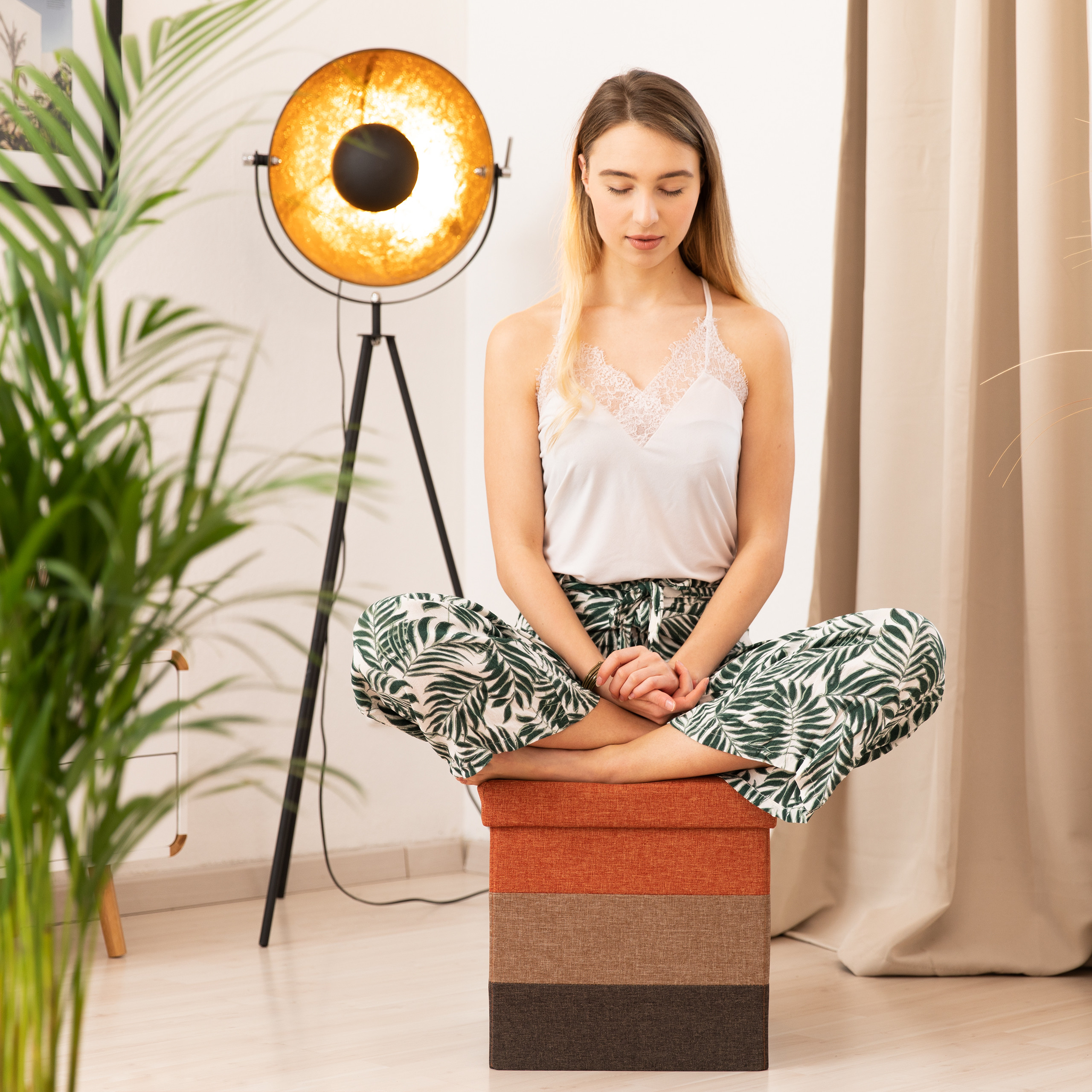 Eine Frau meditiert auf einem orangefarbenen Hocker in einem Raum mit Goldlampe.