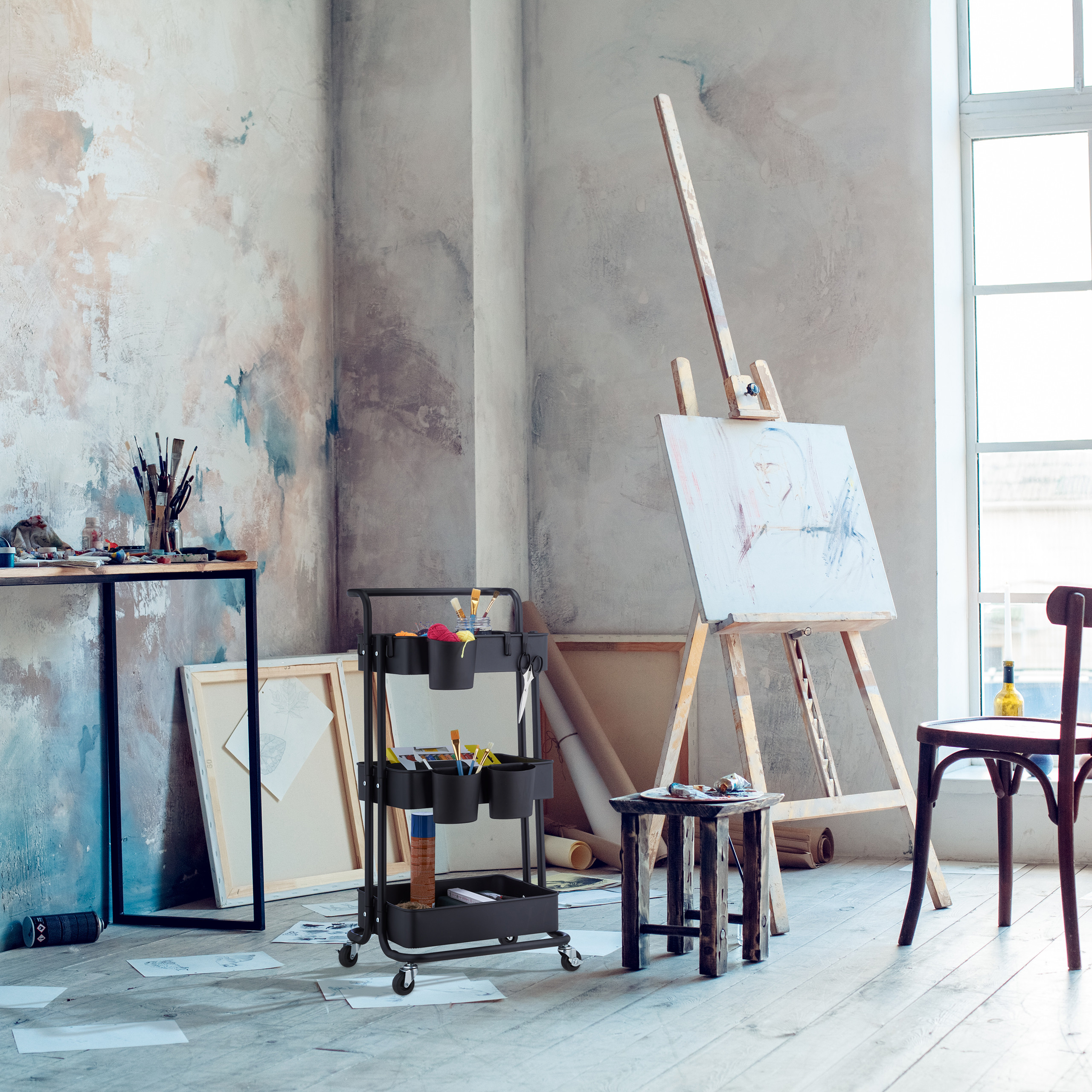 Ein helles Kunststudio mit Staffelei, einem schwarzen Rollwagen für Zubehör und einem Holztisch vor einer grauen Wand.