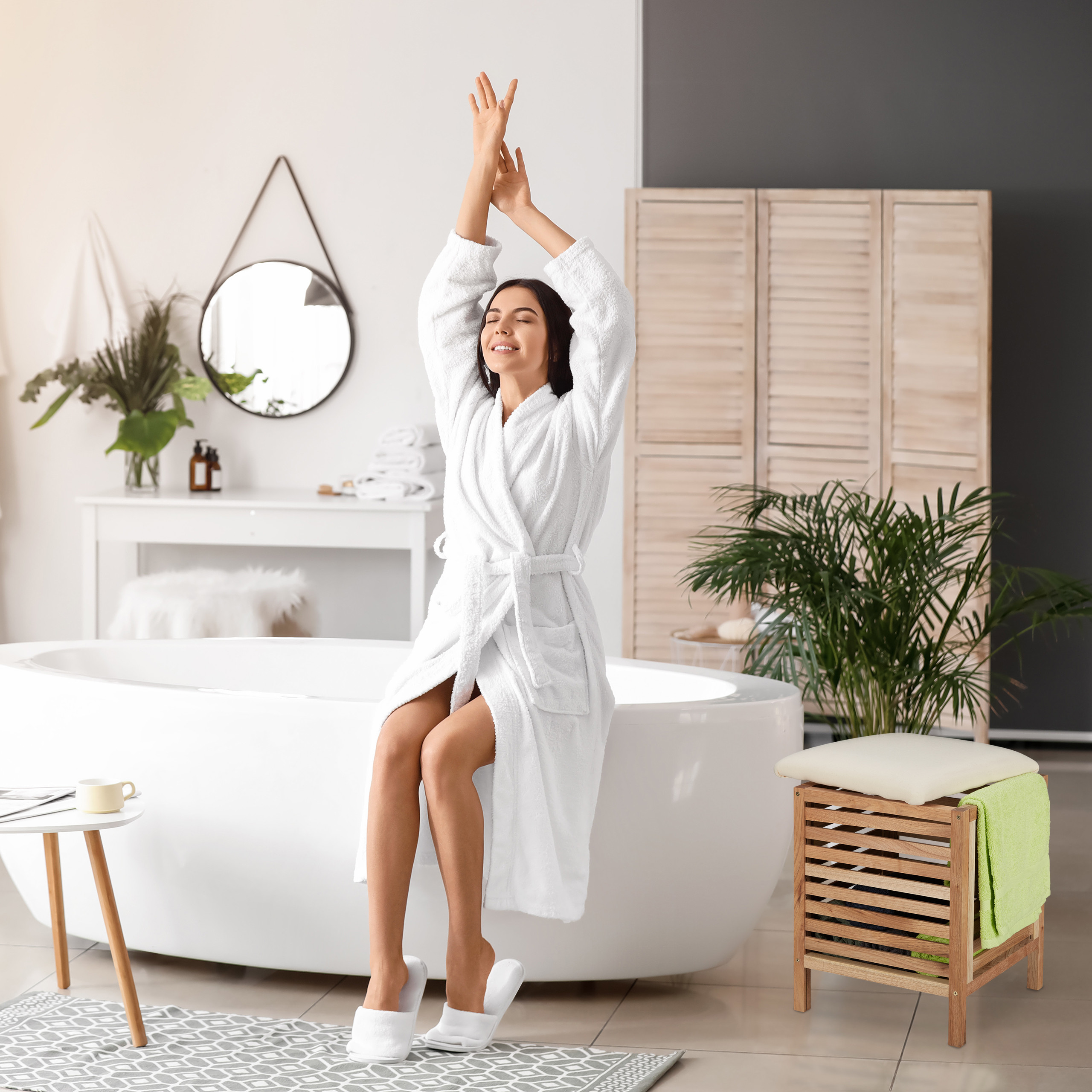 Eine Frau im weißen Bademantel sitzt auf der Badewanne und streckt die Arme in hellem Badezimmer nach oben.