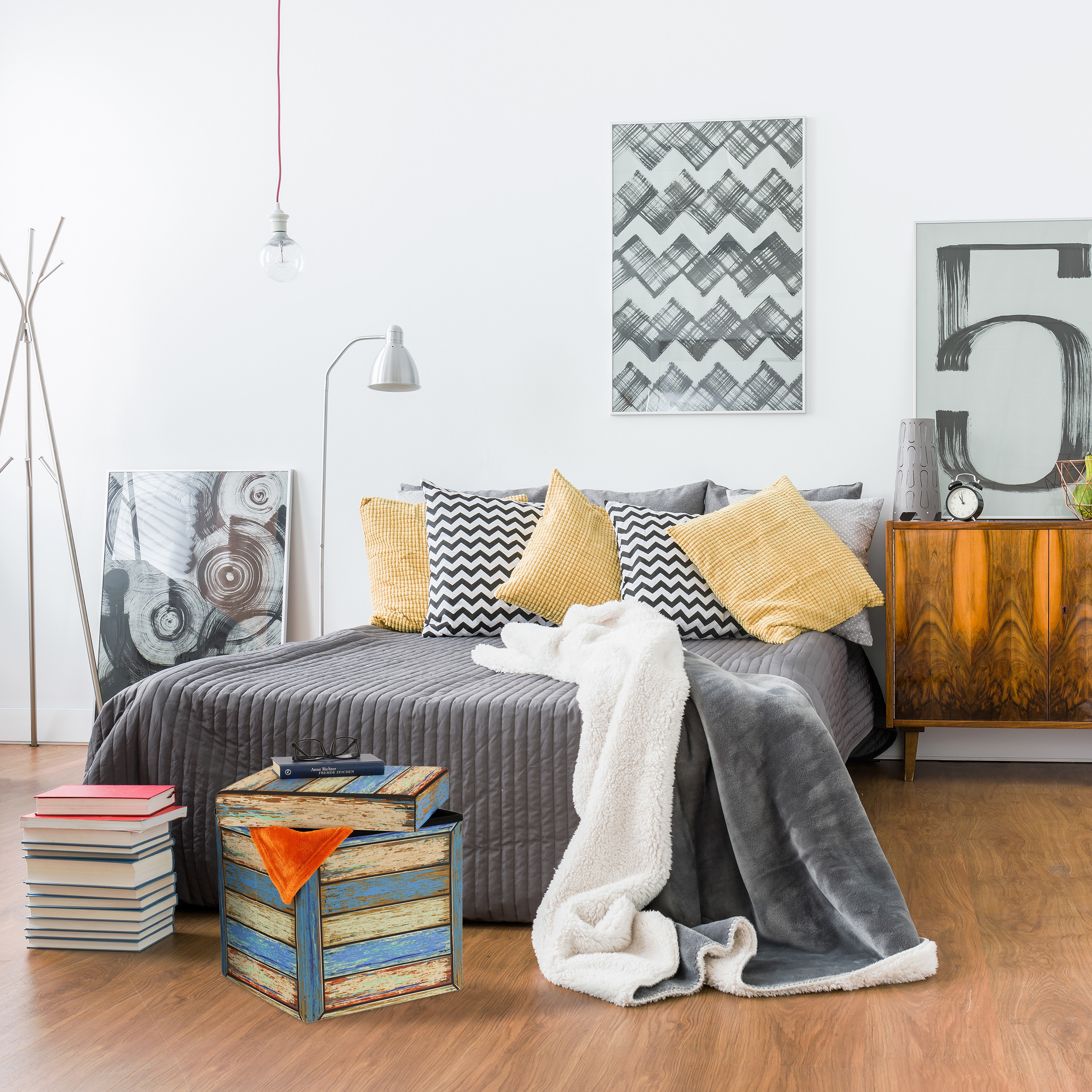 Moderne Schlafzimmer mit grauem Bett, gelben Kissen, Holzkiste und abstrakten Kunstwerken an der Wand.