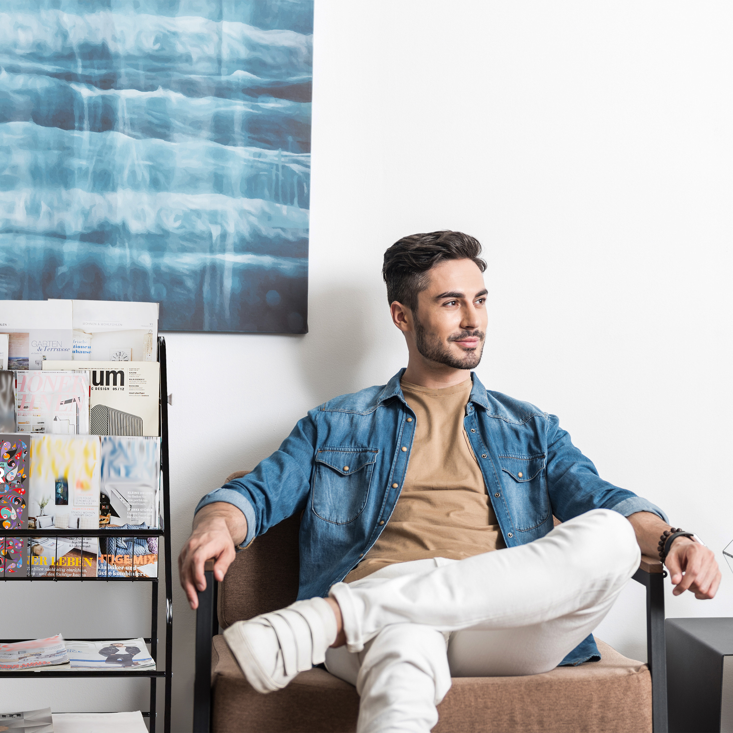 Ein lächelnder Mann sitzt auf einem Stuhl vor einer blauen Leinwand, neben ihm ein Zeitschriftenhalter.