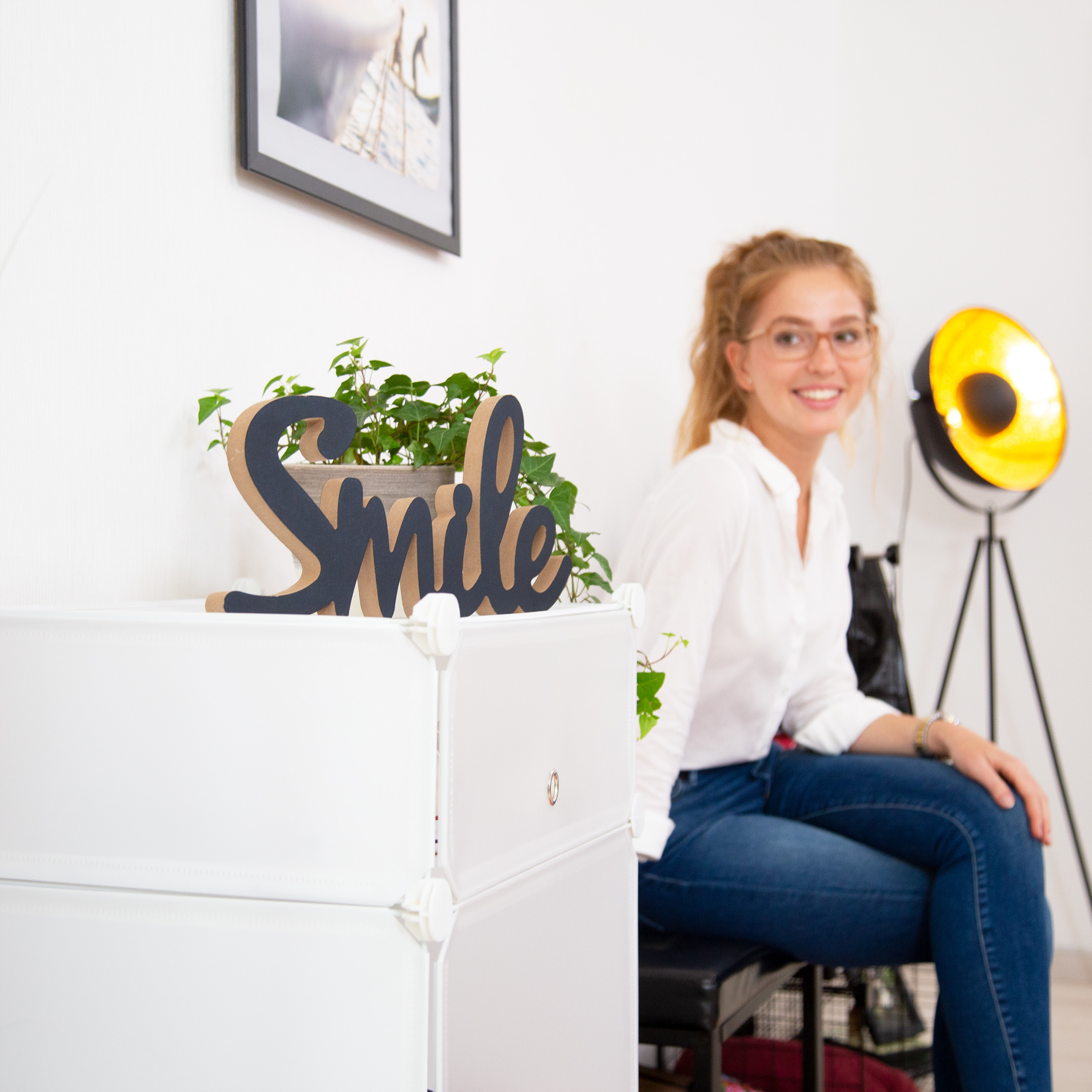 Eine lächelnde Frau sitzt drinnen neben einem weißen Schrank mit einer Pflanze und dem Schriftzug "Smile".
