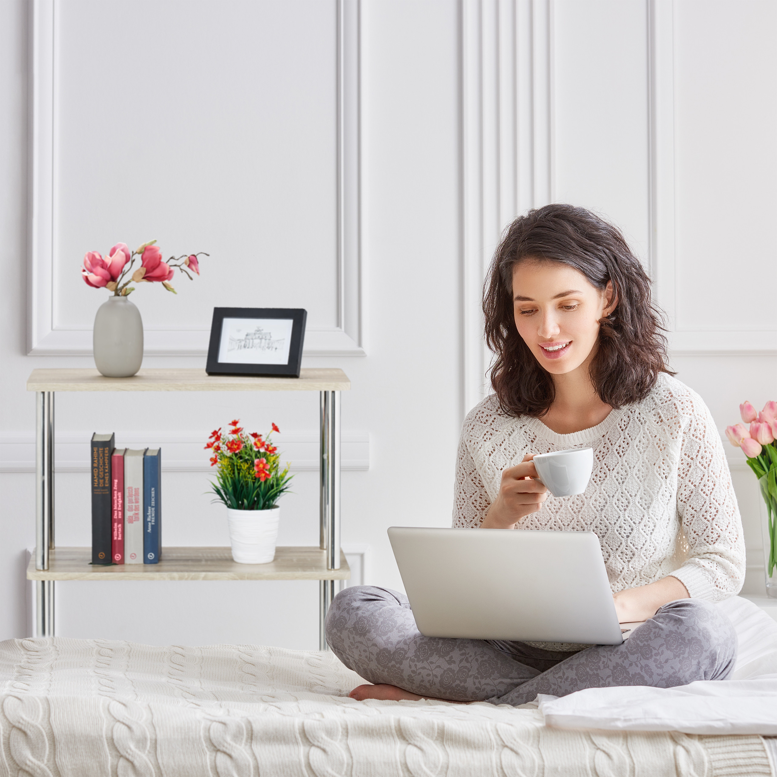 Eine Frau mit Laptop und Kaffeetasse sitzt auf einem Bett in einem hellen Zimmer.