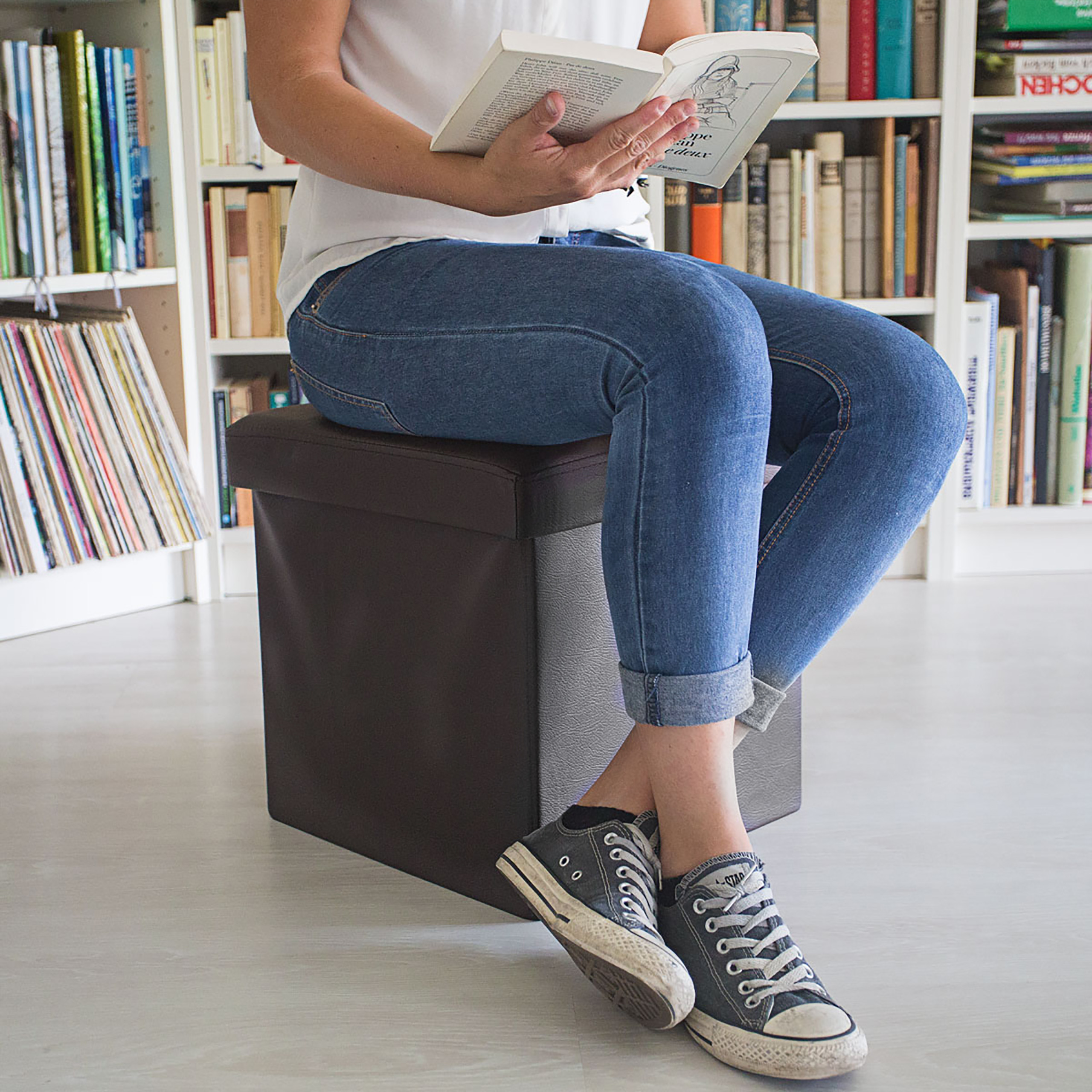 Eine Frau in Jeans und Sneakern sitzt auf einem braunen Hocker vor einem Bücherregal und liest ein Buch.