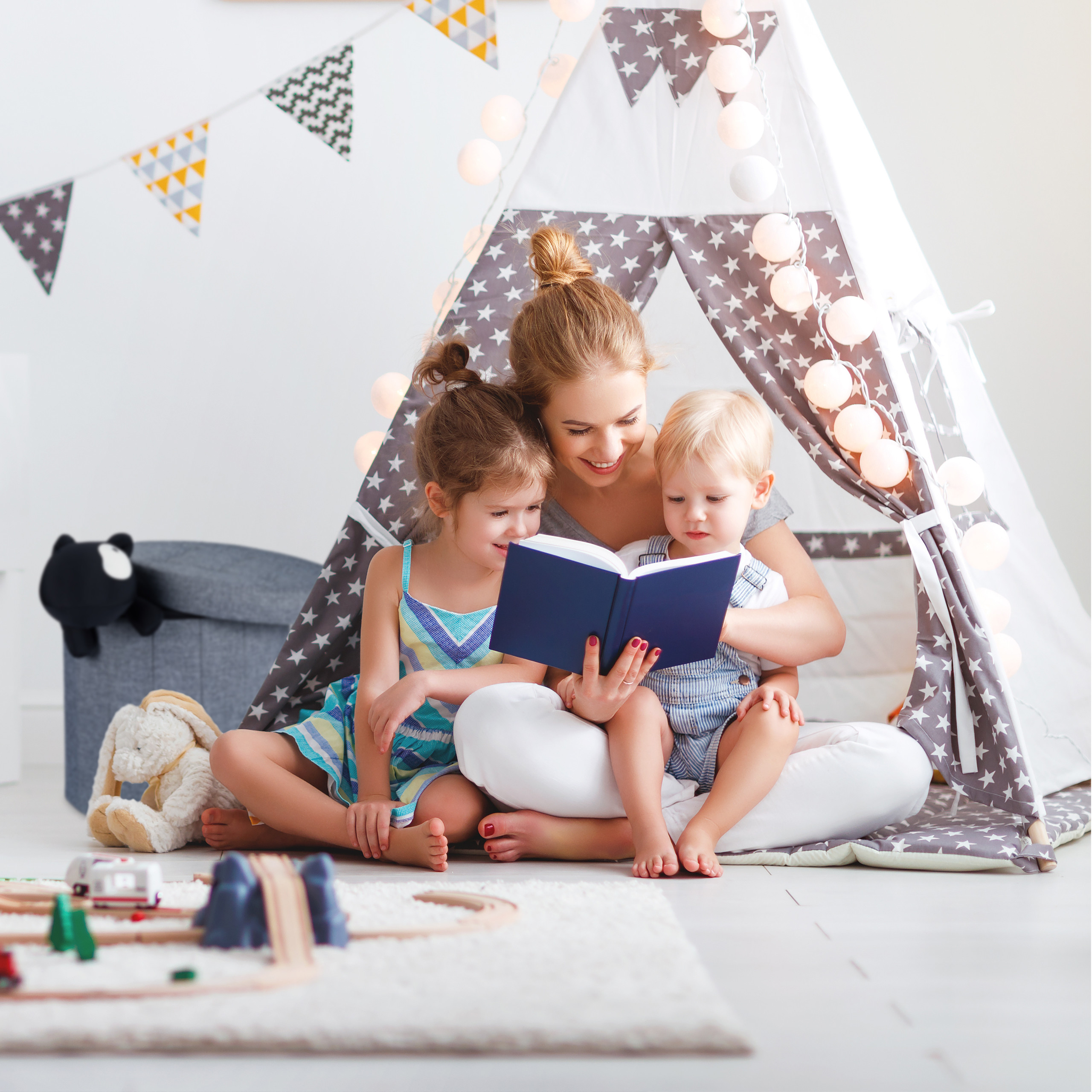Eine Mutter und zwei Kinder lesen ein Buch in einem geschmückten weißen Tipi-Zelt mit Lichterkette in einem hellen Raum.