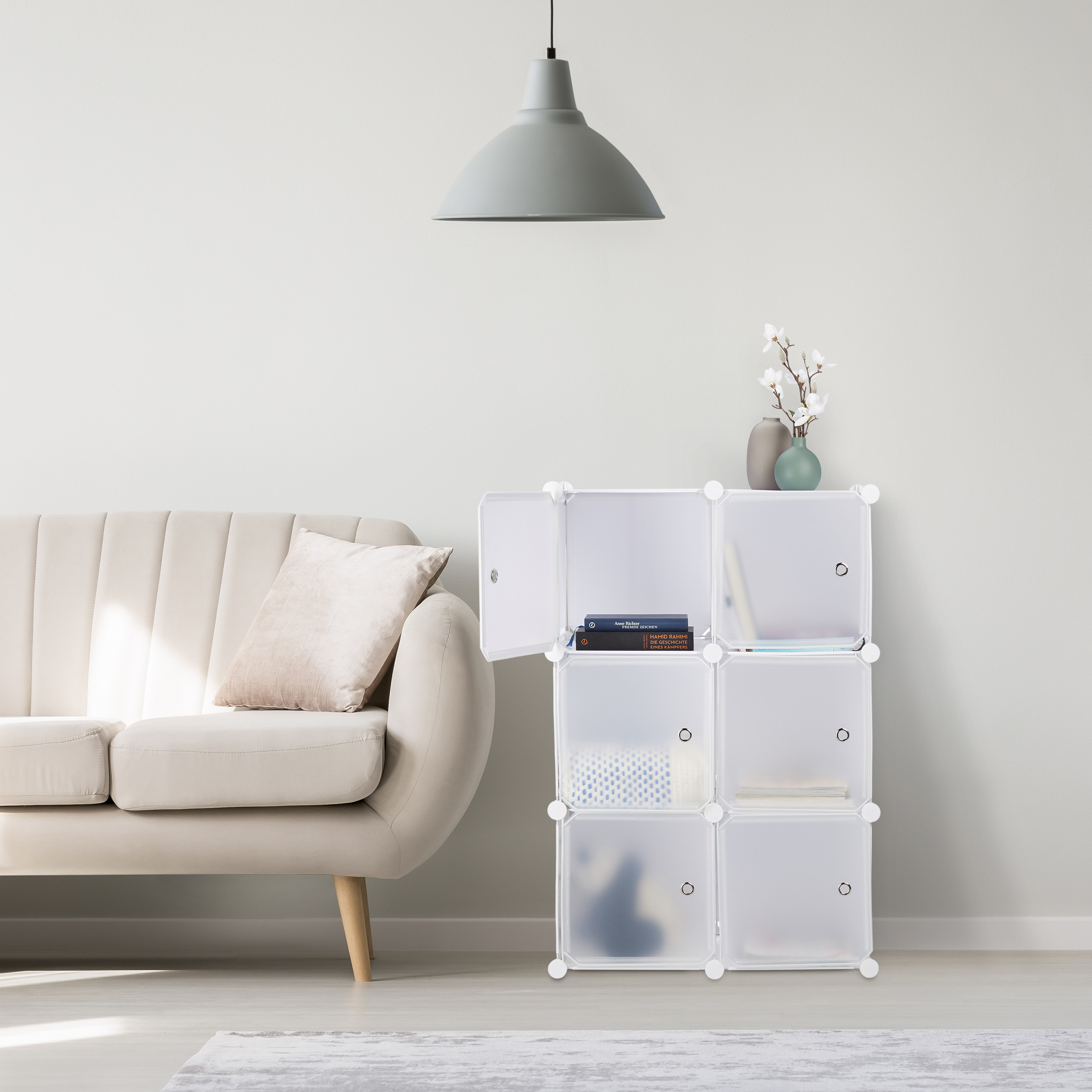 Ein heller Wohnraum mit beigem Sofa, grauer Hängelampe und weißem Kunststoff-Regalsystem mit Büchern.