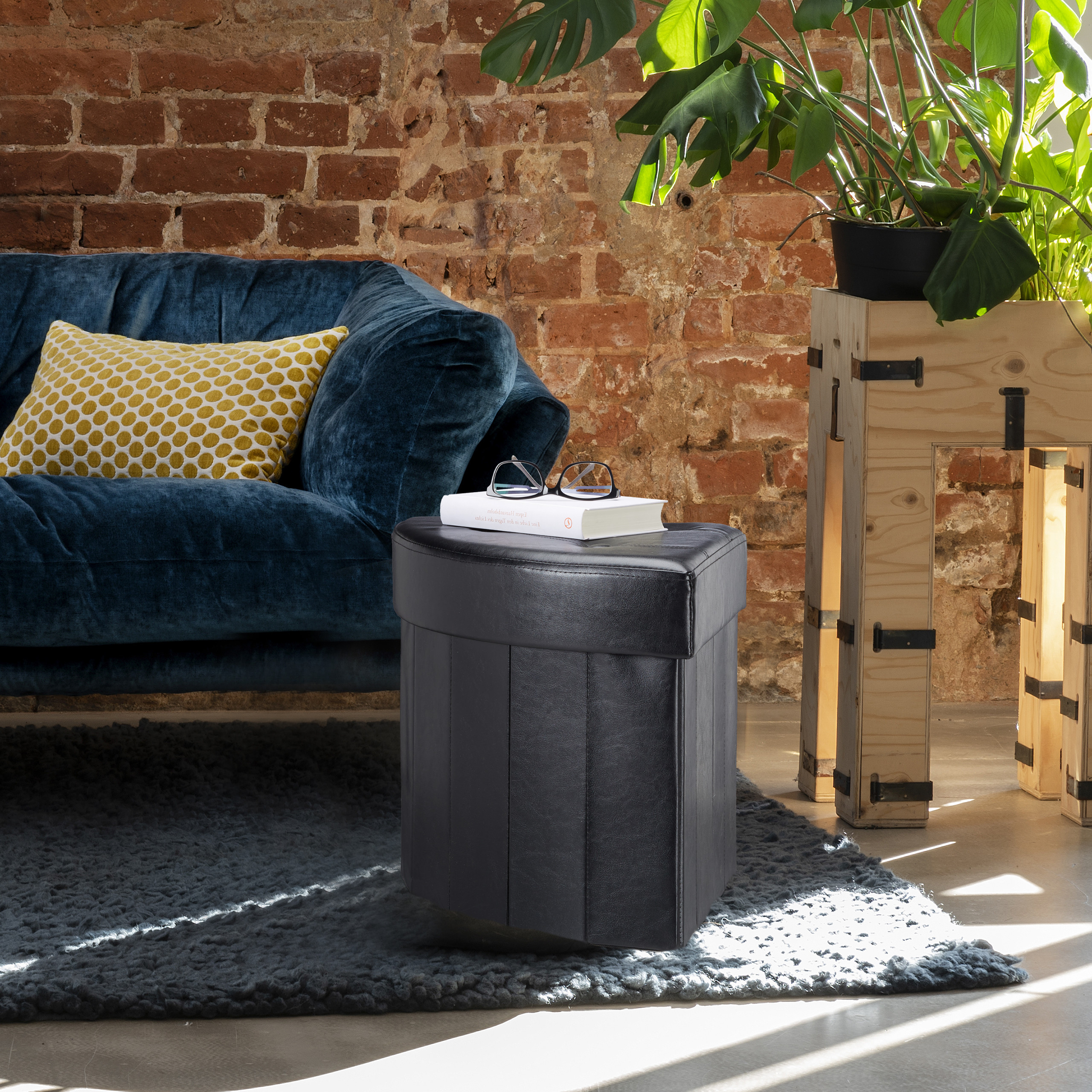Ein schwarzer Hocker auf einem Teppich vor einem blauen Sofa und einer Ziegelwand.