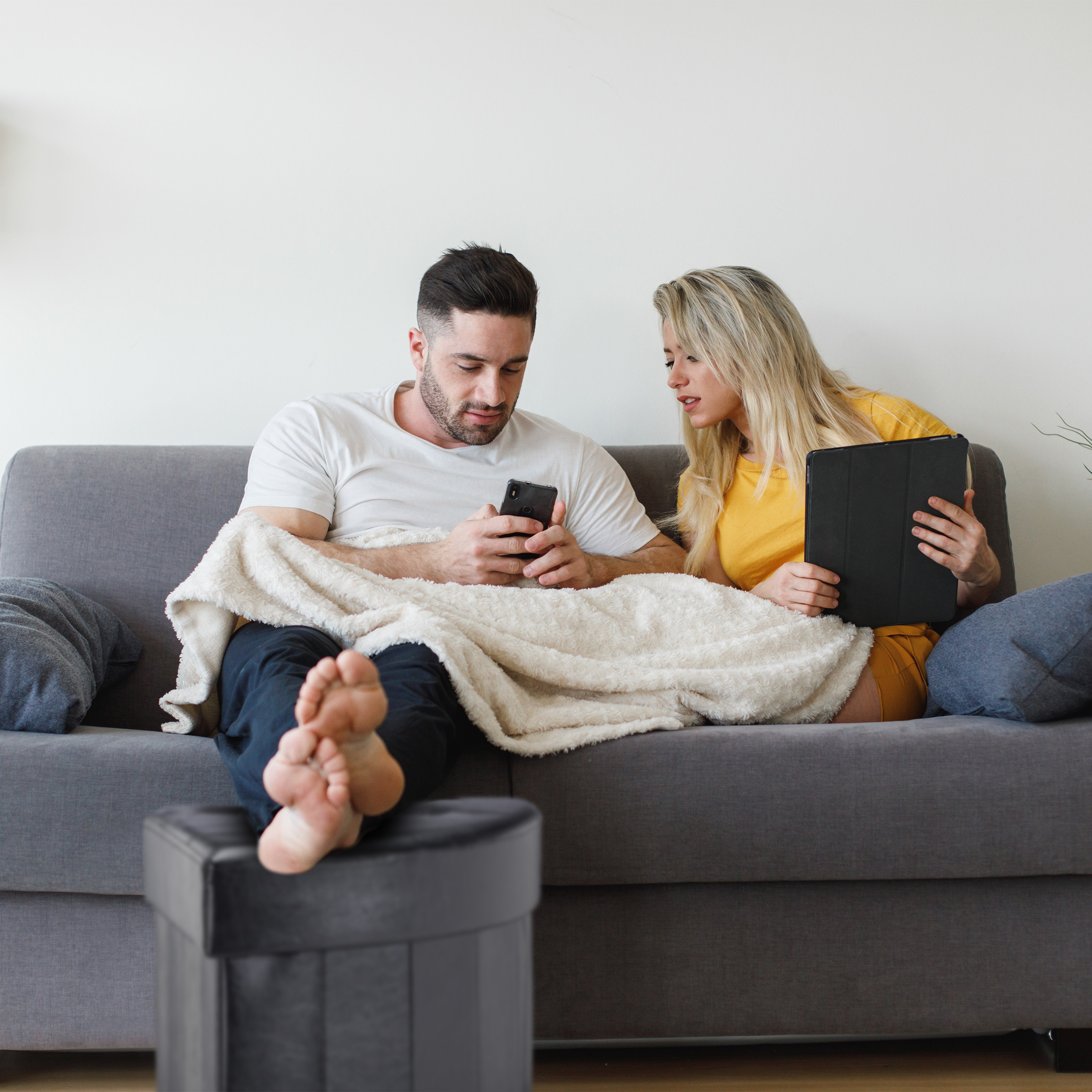 Ein Mann und eine Frau entspannen auf einem grauen Sofa und schauen auf ein Handy und ein Tablet.