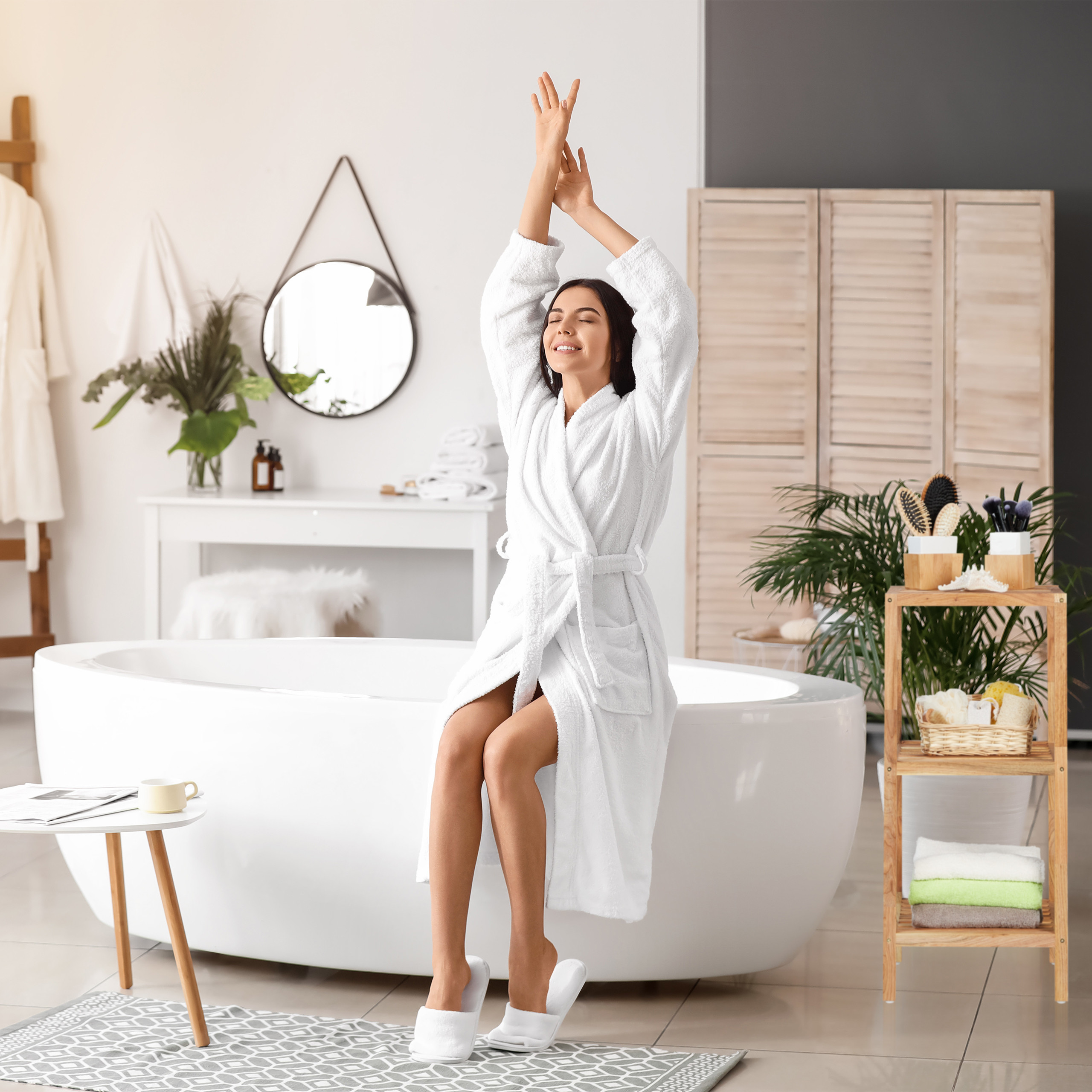 Frau im weißen Bademantel sitzt auf einer Badewanne und streckt sich mit geschlossenen Augen.