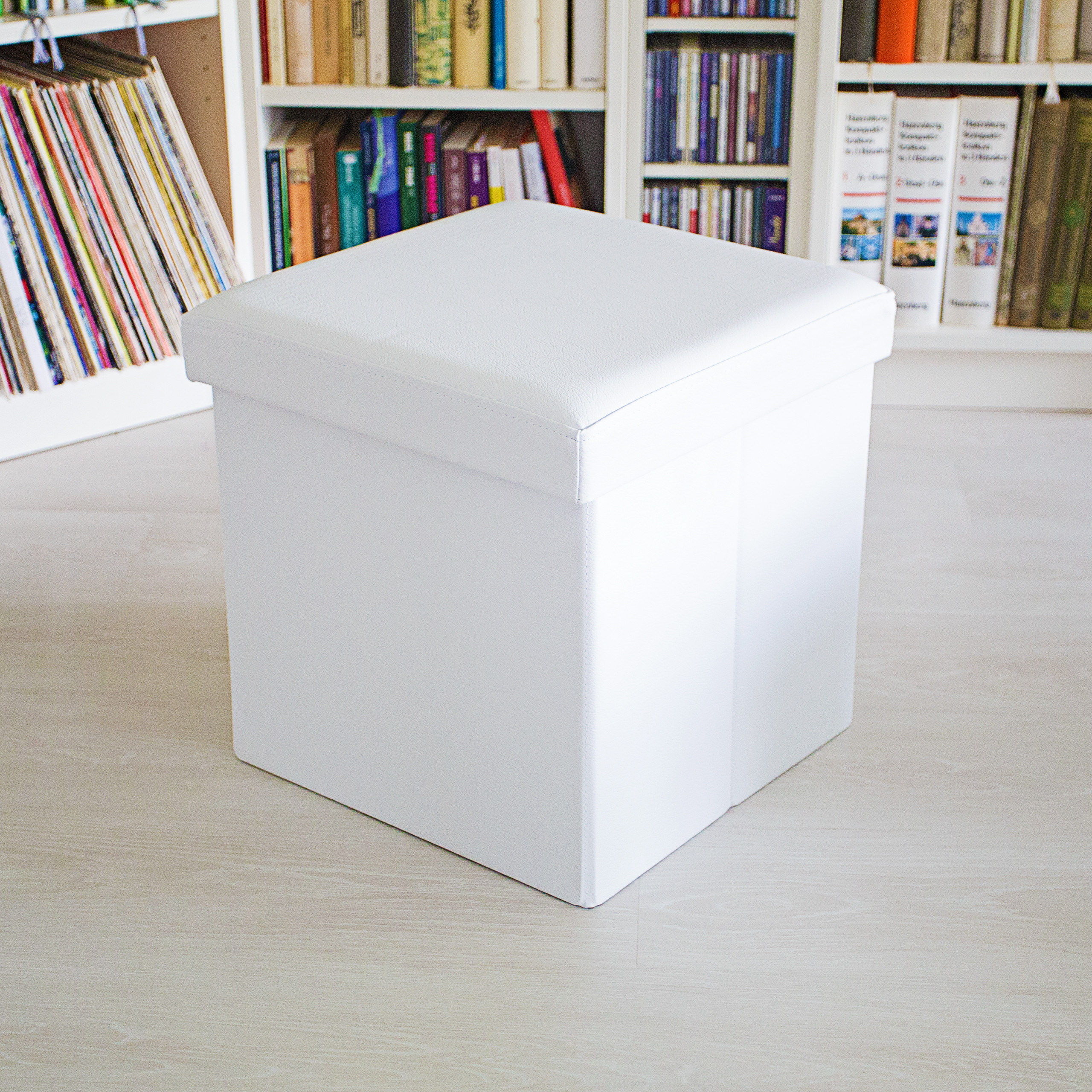Ein weißer, quadratischer Sitzhocker steht vor hellen Bücherregalen.