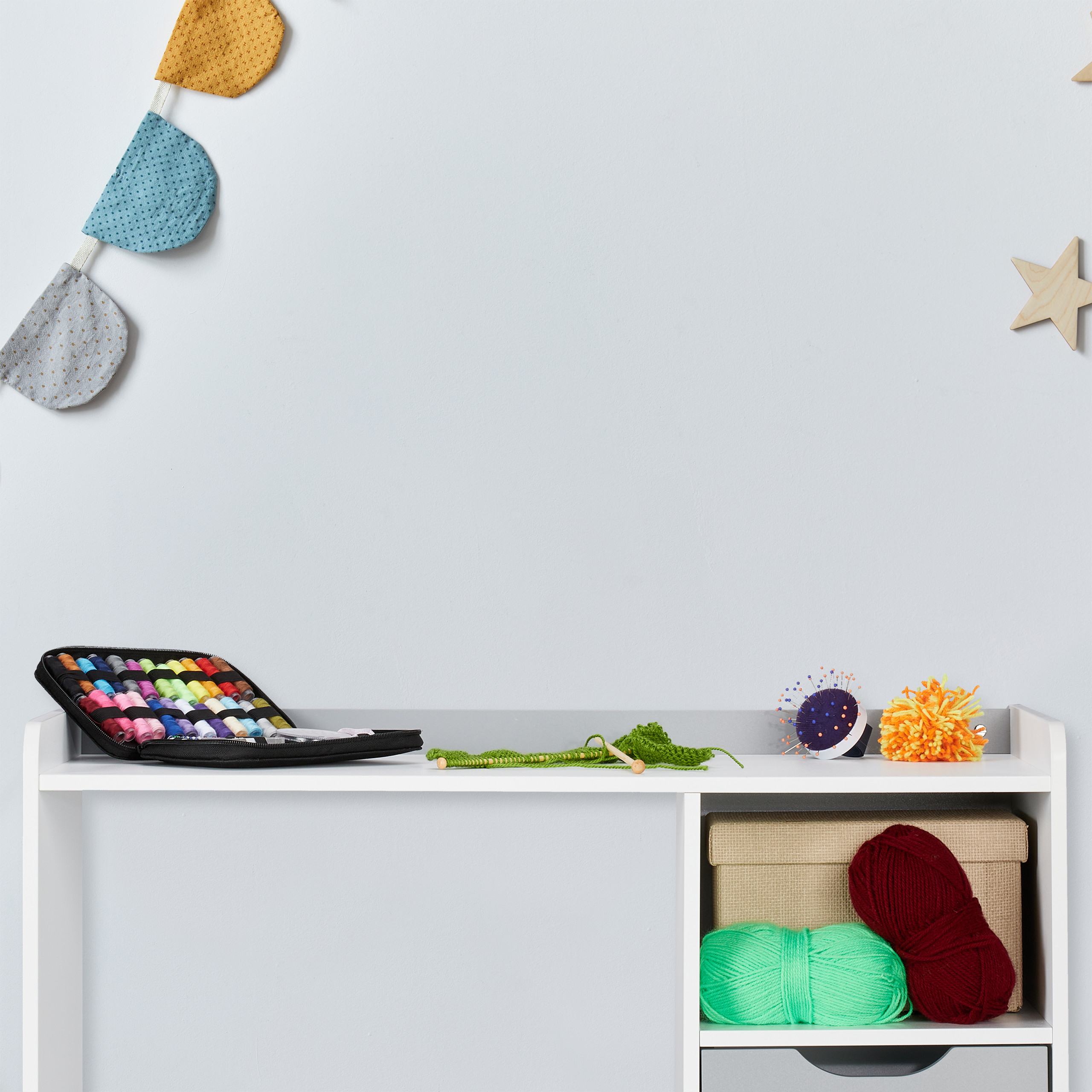 Ein weißes Regal mit Nähutensilien, Garn, Strickzeug und bunten Deko-Fähnchen an der grauen Wand.