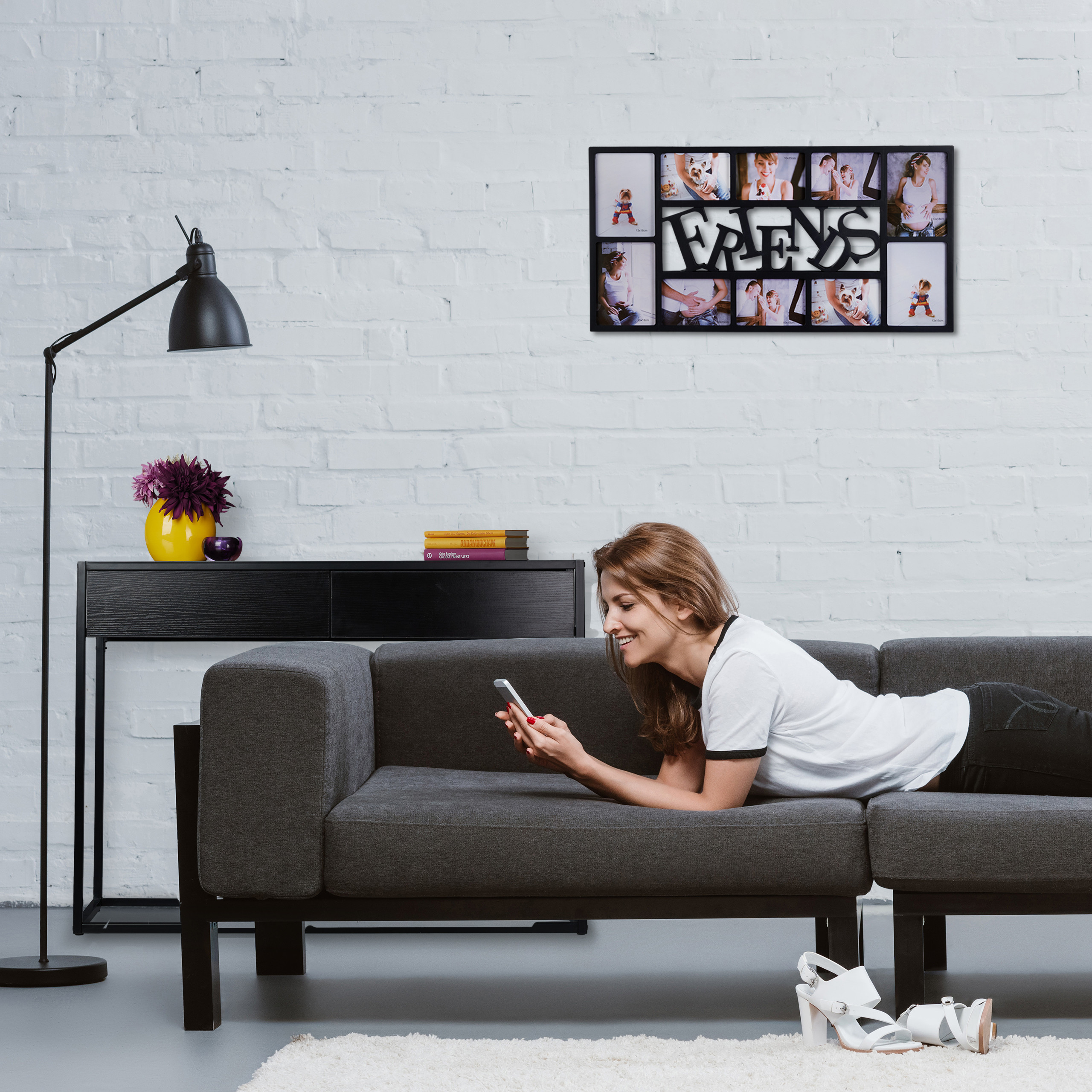 Eine Frau liegt lächelnd auf einem grauen Sofa und nutzt ihr Smartphone. An der weißen Backsteinwand hängt ein Fotorahmen.